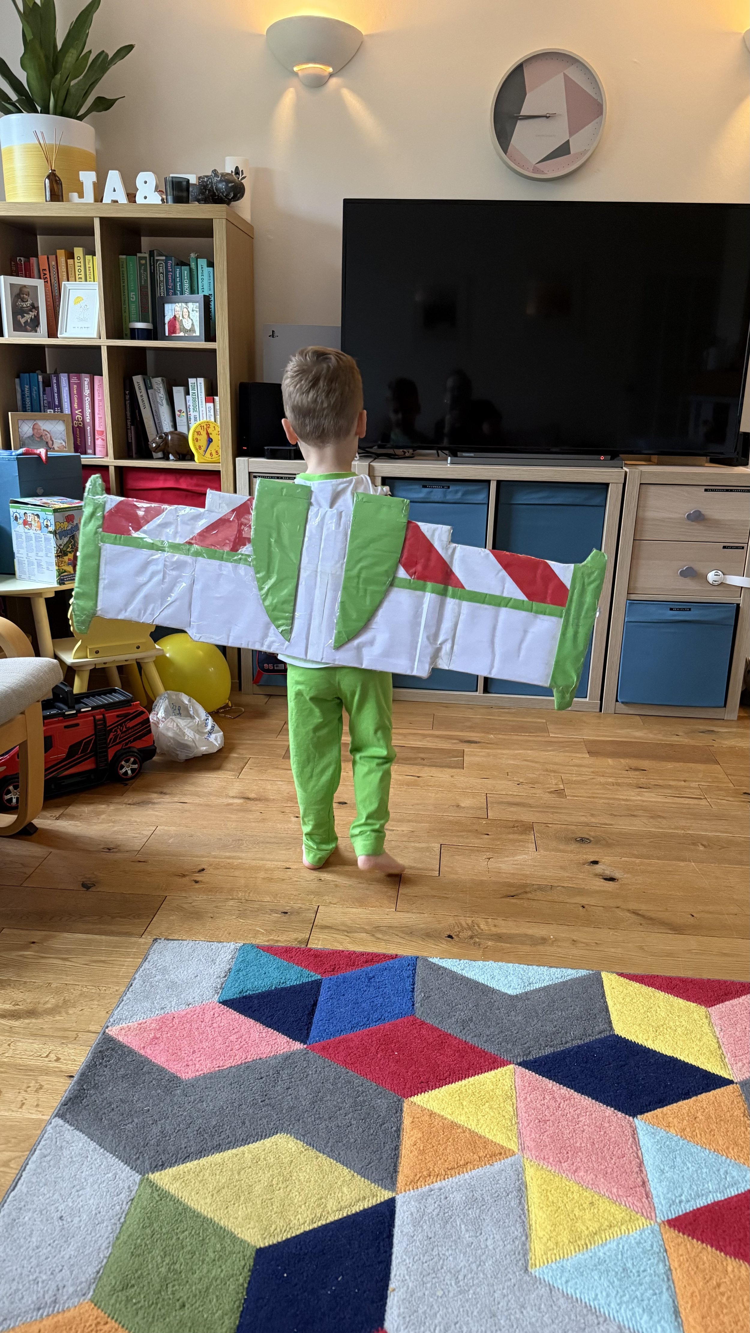 A young boy dressed in a green costume with large wings stands in a living room facing a large flat-screen TV. The room has a bamboo cabinet filled with books and framed photos, a colorful geometric rug, and various toys on the floor.