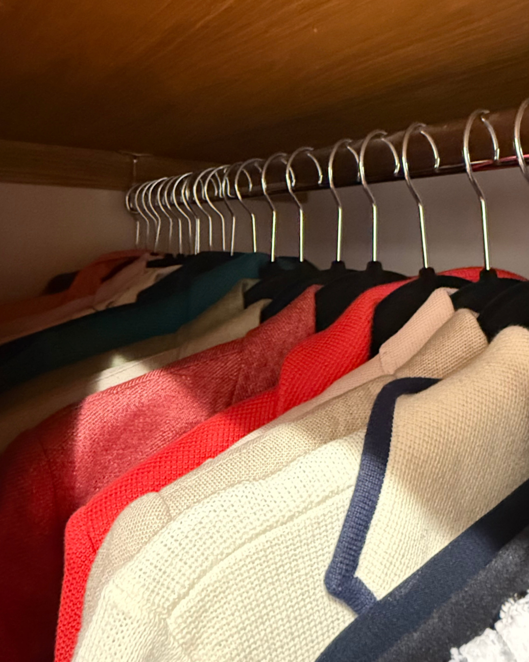 A wardrobe with a wooden shelf holding hangers with clothes like sweaters and shirts in various colors including red, black, beige, and teal.