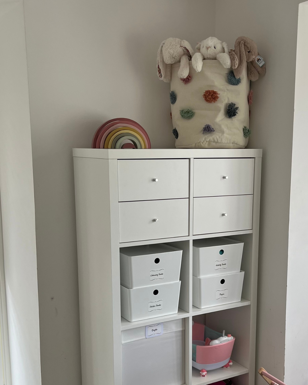 A white storage unit with four small drawers on top, four labeled fabric bins in the middle, and open shelves at the bottom, topped with a decorative rainbow and a hanging plush toy bag filled with stuffed animals.
