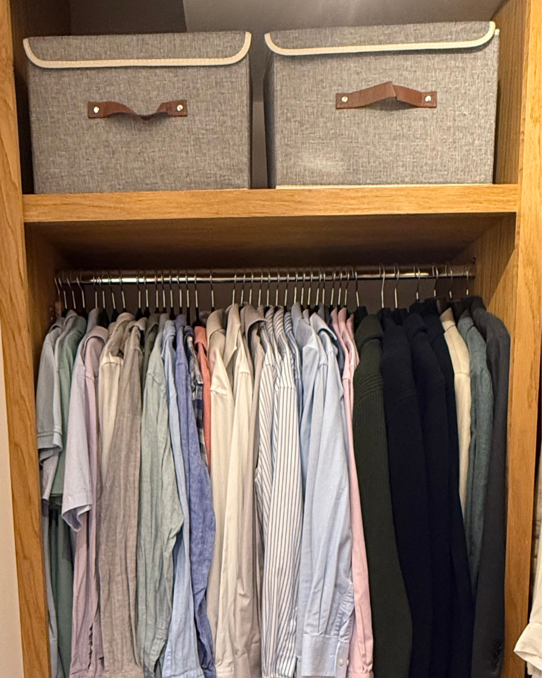 A wooden closet with a row of hanging shirts of various colors and styles, and two fabric storage bins on the top shelf with leather handles.