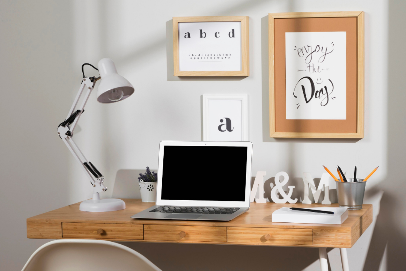 Home office desk with a white lamp, laptop, decorative letters 'M&M', a notebook with a pen, a plant, and framed quotes on the wall reading 'a b c d' and 'enjoy the day'.