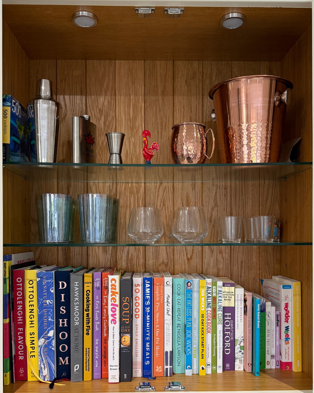A wooden bookshelf with three shelves containing glassware, cocktail shakers, a copper bucket, and a row of cookbooks.