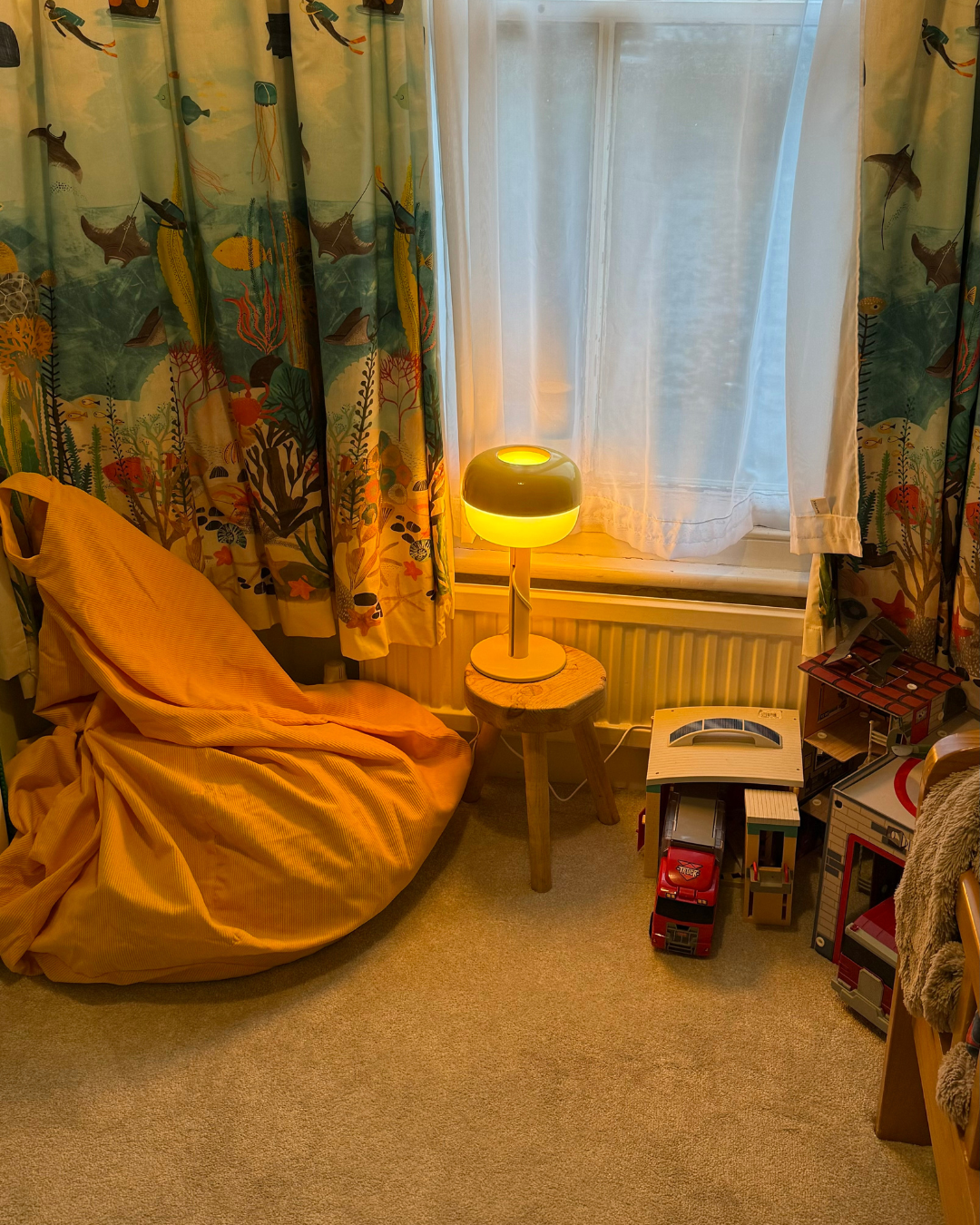 A cozy corner of a child's room with a bean bag chair covered in orange fabric, a small wooden stool holding a modern lamp with a glowing yellow-green light, a window with white curtains and colorful ocean-themed curtains, and toy cars and building b