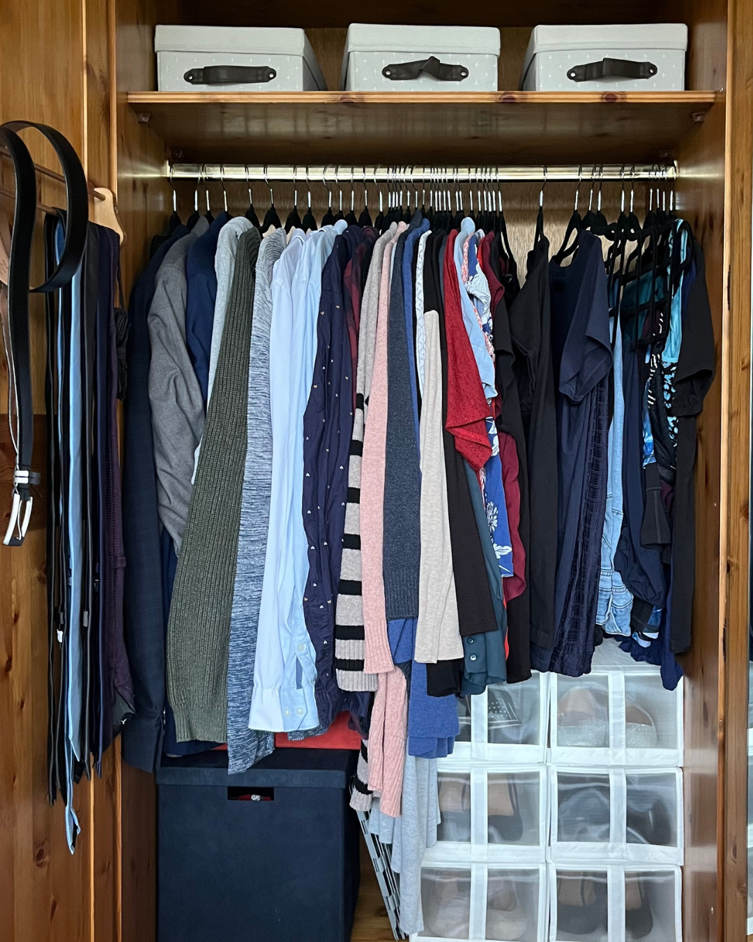 A wooden closet filled with men's shirts, jackets, and workout clothes on hangers, with storage boxes on top and plastic drawers at the bottom.