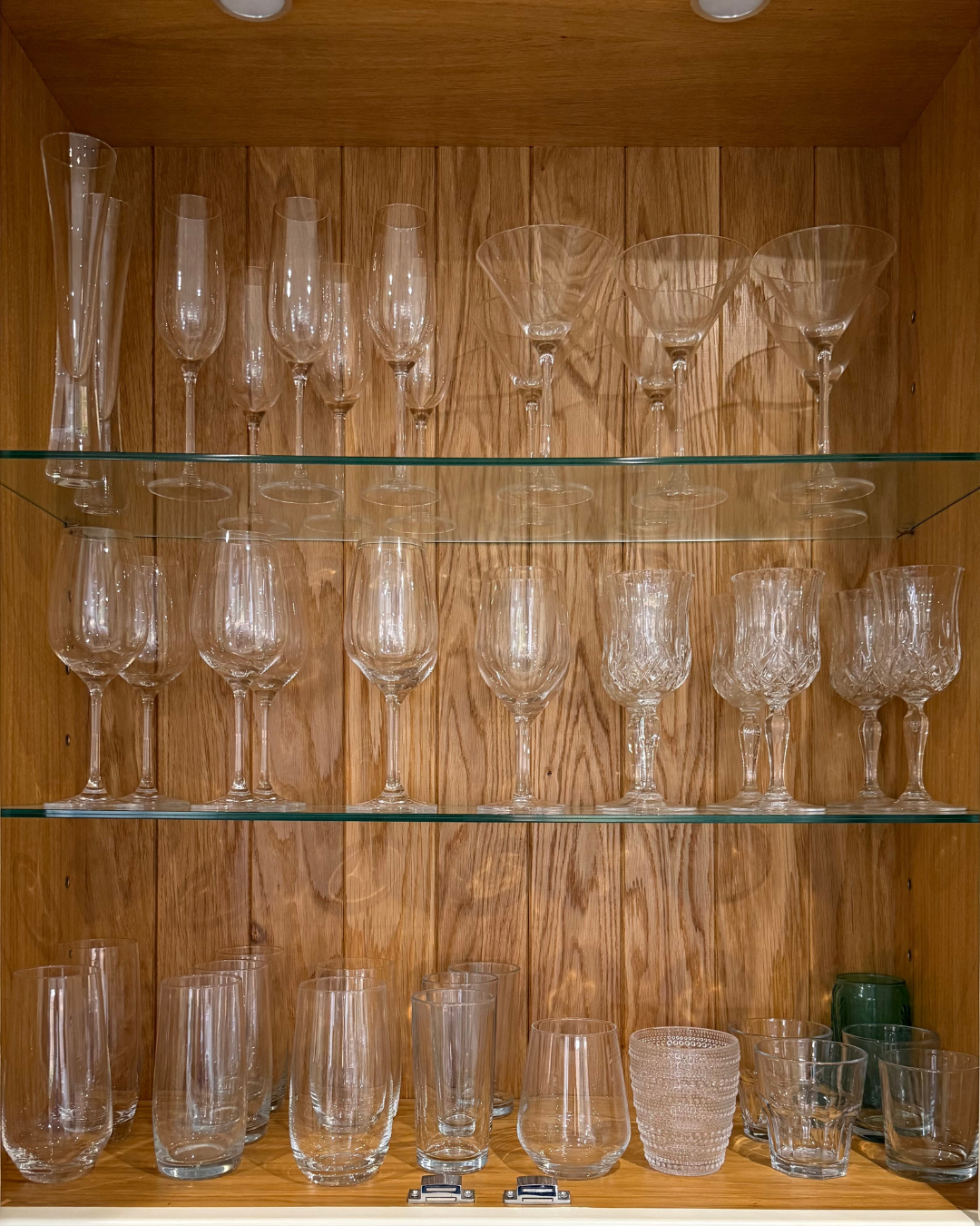 Glassware collection in a wooden cabinet with three shelves, containing champagne flutes, martini glasses, wine glasses, tumblers, and a decorative glass.