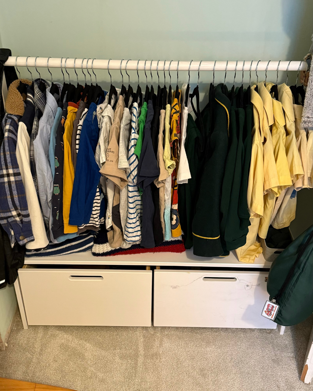 Closet with hanging assorted childhood shirts, jackets, and sweaters, and white storage drawers below.