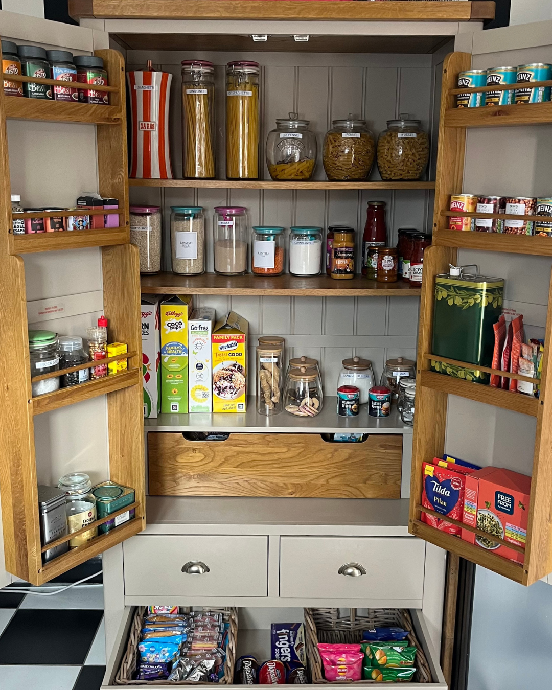 Open pantry with shelves holding jars of pasta, rice, beans, and canned goods, along with boxes of cereal and snack foods in baskets, on a black and white checkered floor.
