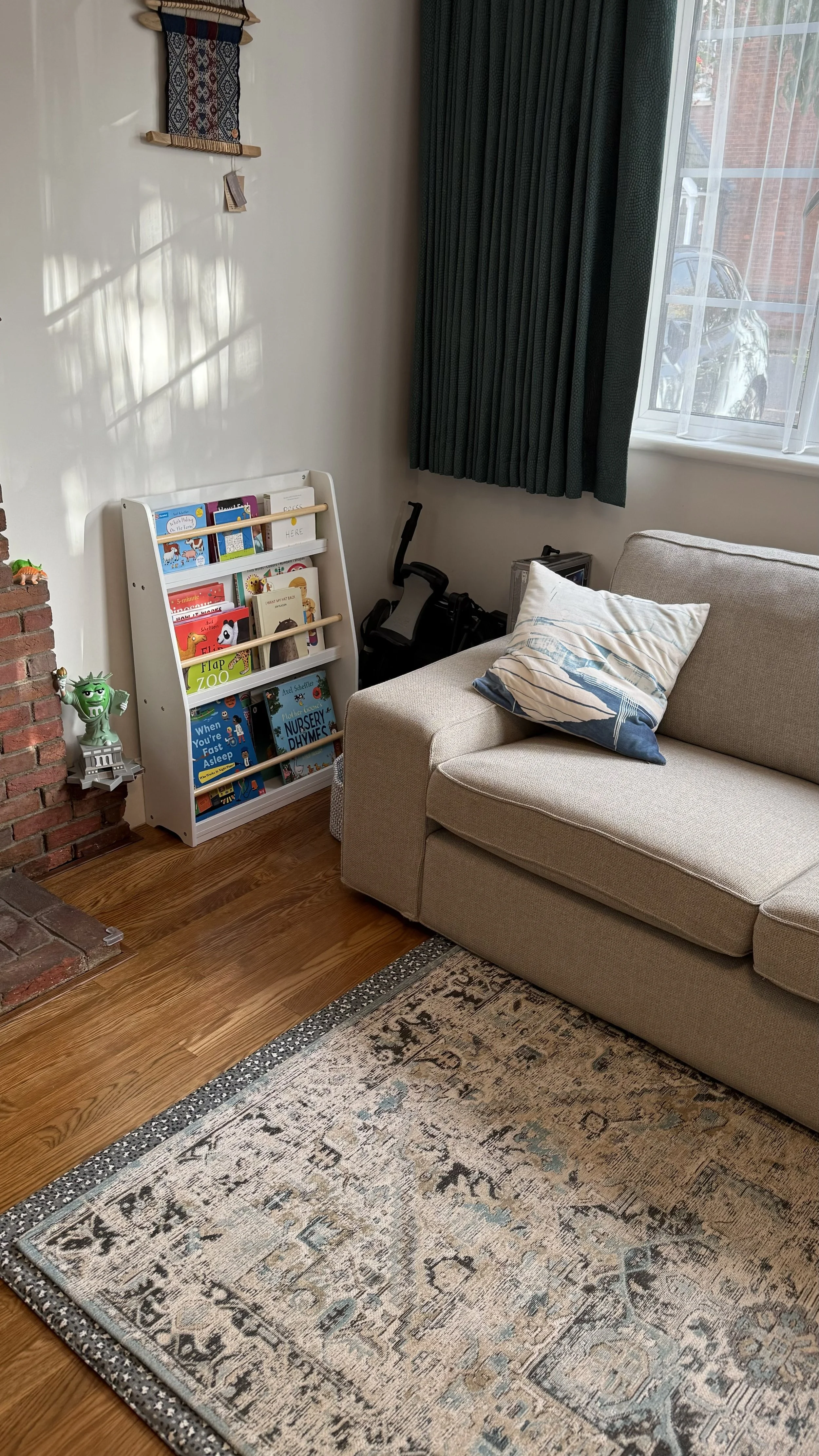 A cozy living room corner with a beige sofa, a decorative pillow, and a window with teal curtains. There is a small white bookshelf filled with children's books, a green toy figure on a small shelf, and a treadmill behind the sofa. The room has hardw