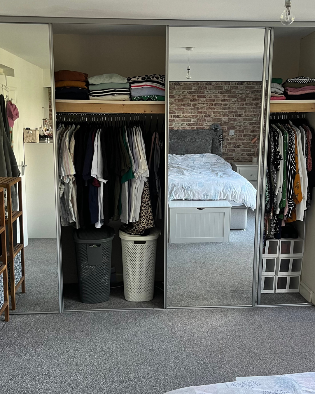A bedroom with a large mirrored wardrobe containing hanging clothes and shelves with folded clothes, a bed with a white duvet, a brick accent wall, and a white nightstand.