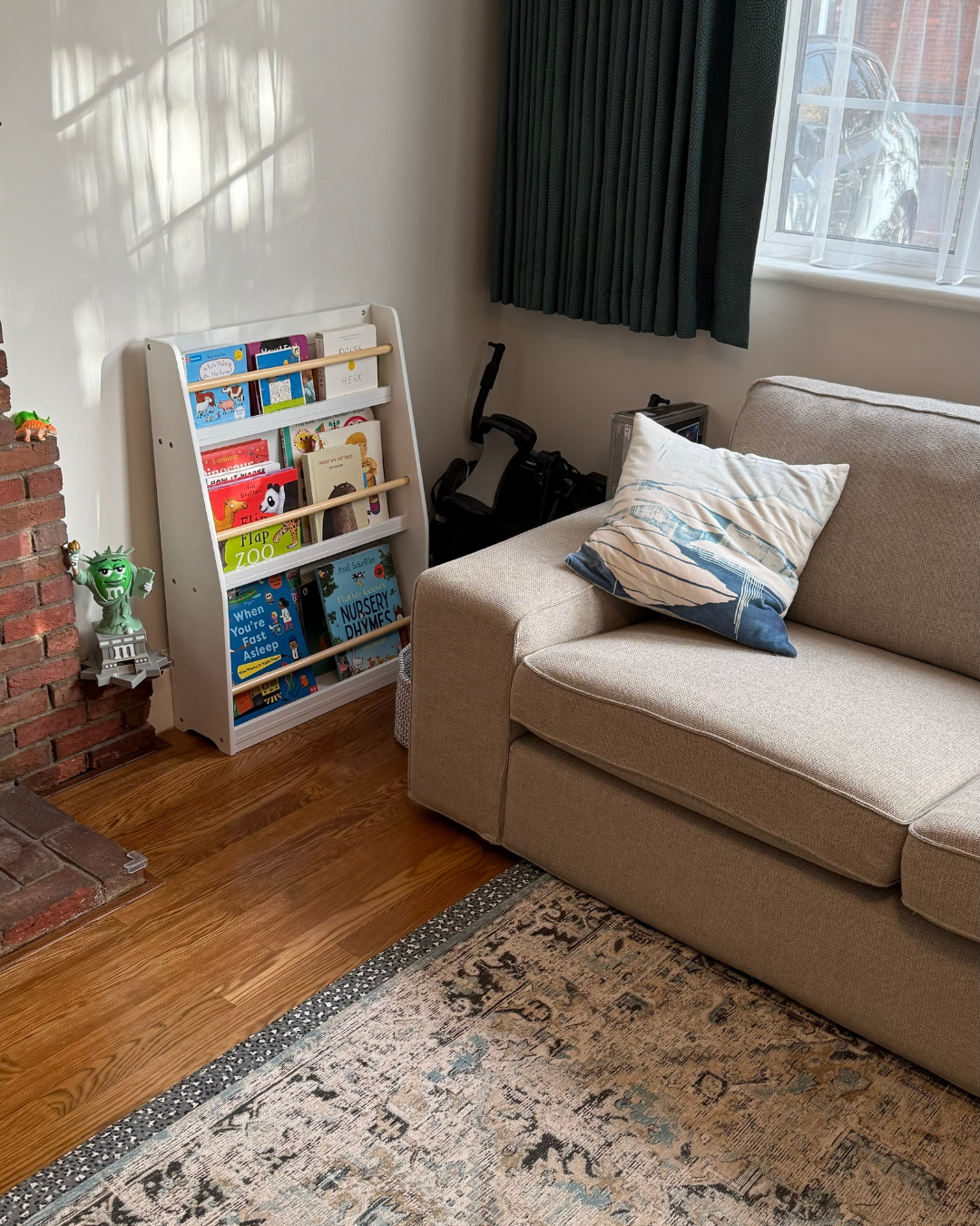A cozy living room corner with a beige sofa, a striped pillow, a bookshelf filled with children's books, a black exercise bike behind the sofa, a window with dark curtains, a brick fireplace with a small toy on the mantel, and a patterned area rug on