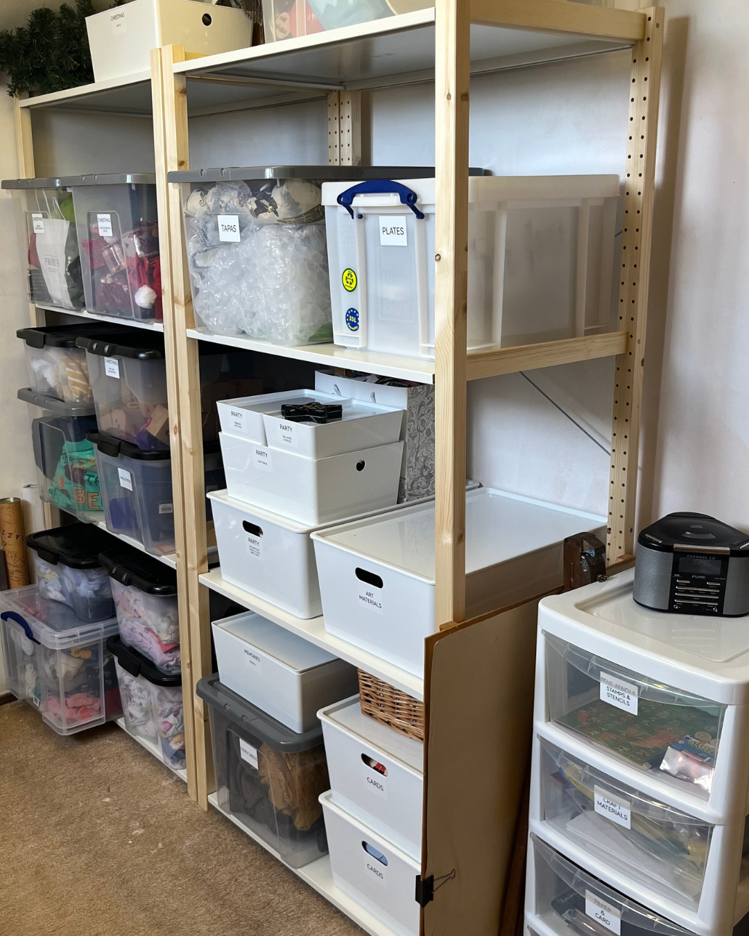 Shelving unit with labeled plastic storage bins containing craft supplies, art materials, and decorations, in a craft or storage room.