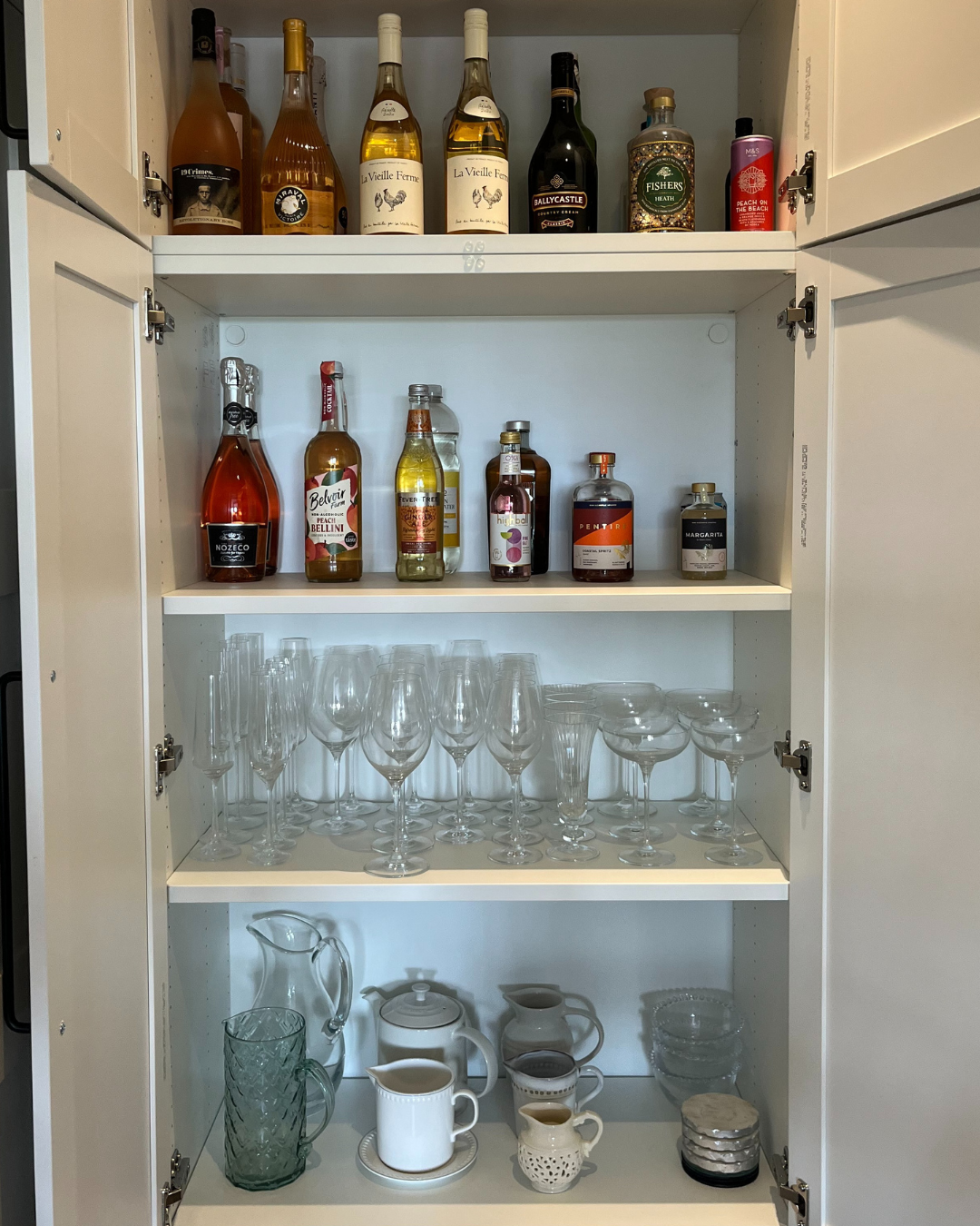 Cabinet with alcohol bottles on top, liquor bottles on middle shelf, wine glasses on the bottom shelf, and glassware and teapots on the floor.