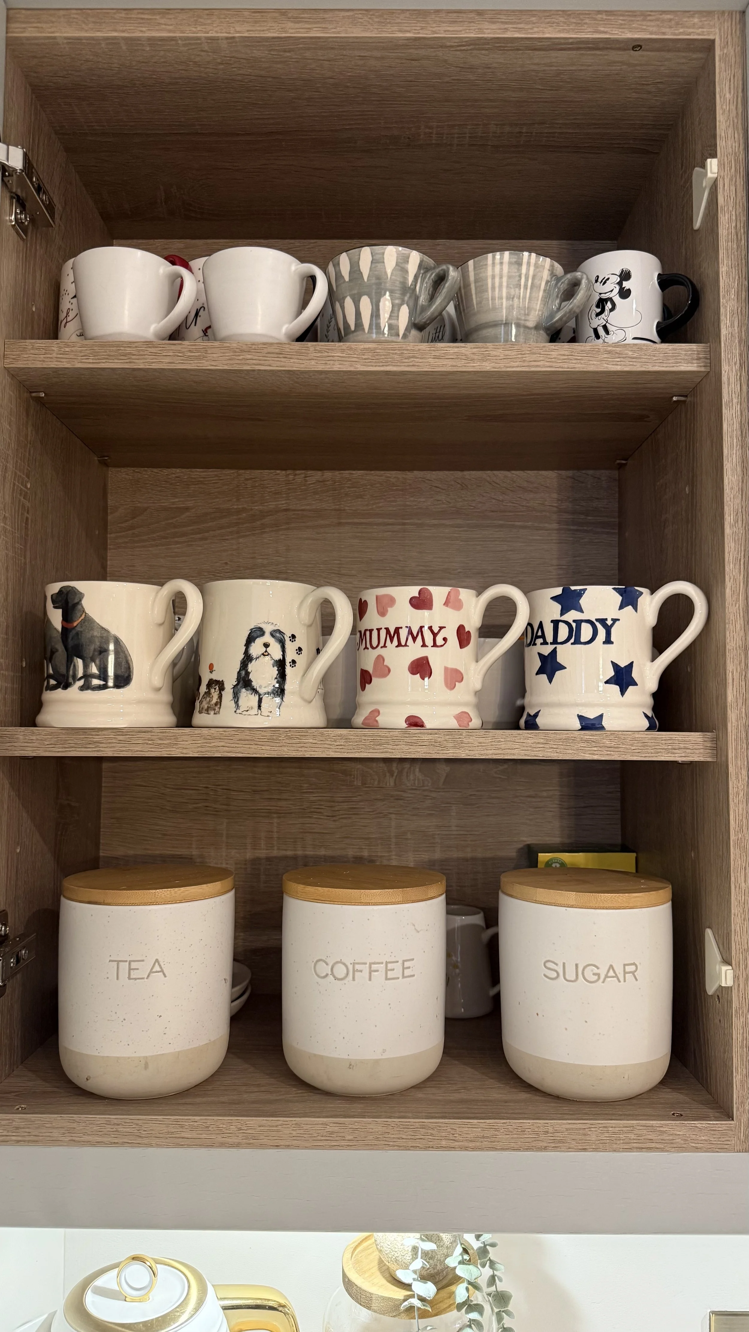 Wooden kitchen cabinet with three shelves. The top shelf has white and patterned mugs, including a Mickey Mouse mug. The middle shelf has four mugs with dog and heart-themed designs, including one labeled 'MUMMY' and another labeled 'DADDY.' The bott