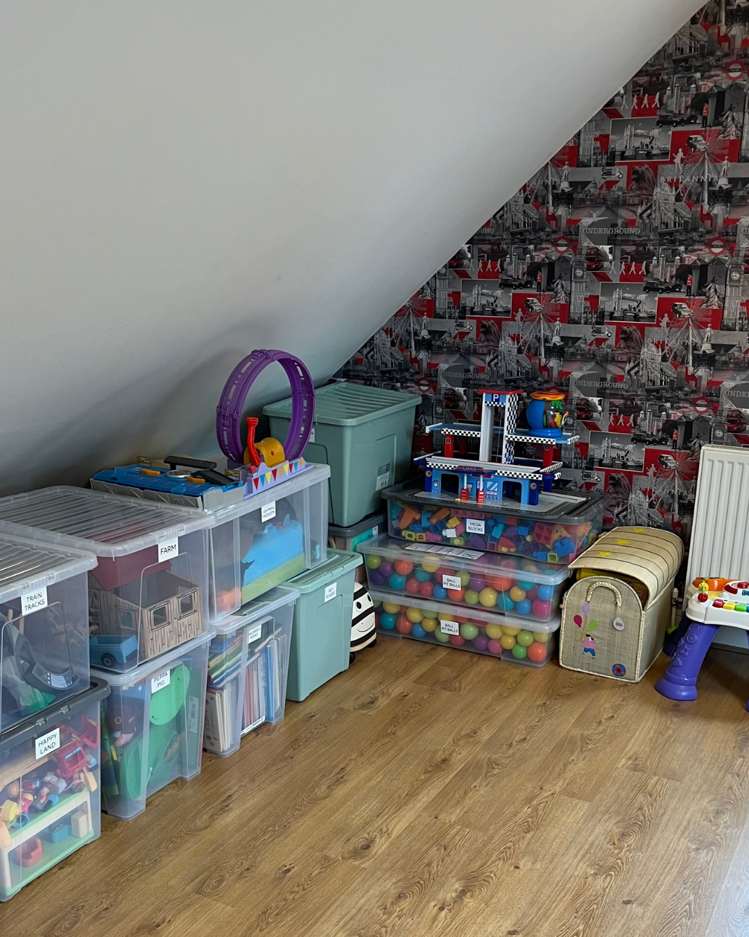 Organised corner of a converted loft with plastic storage bins filled with toys, a purple toy racetrack, a blue garage playset, and a wallpaper featuring London landmarks.