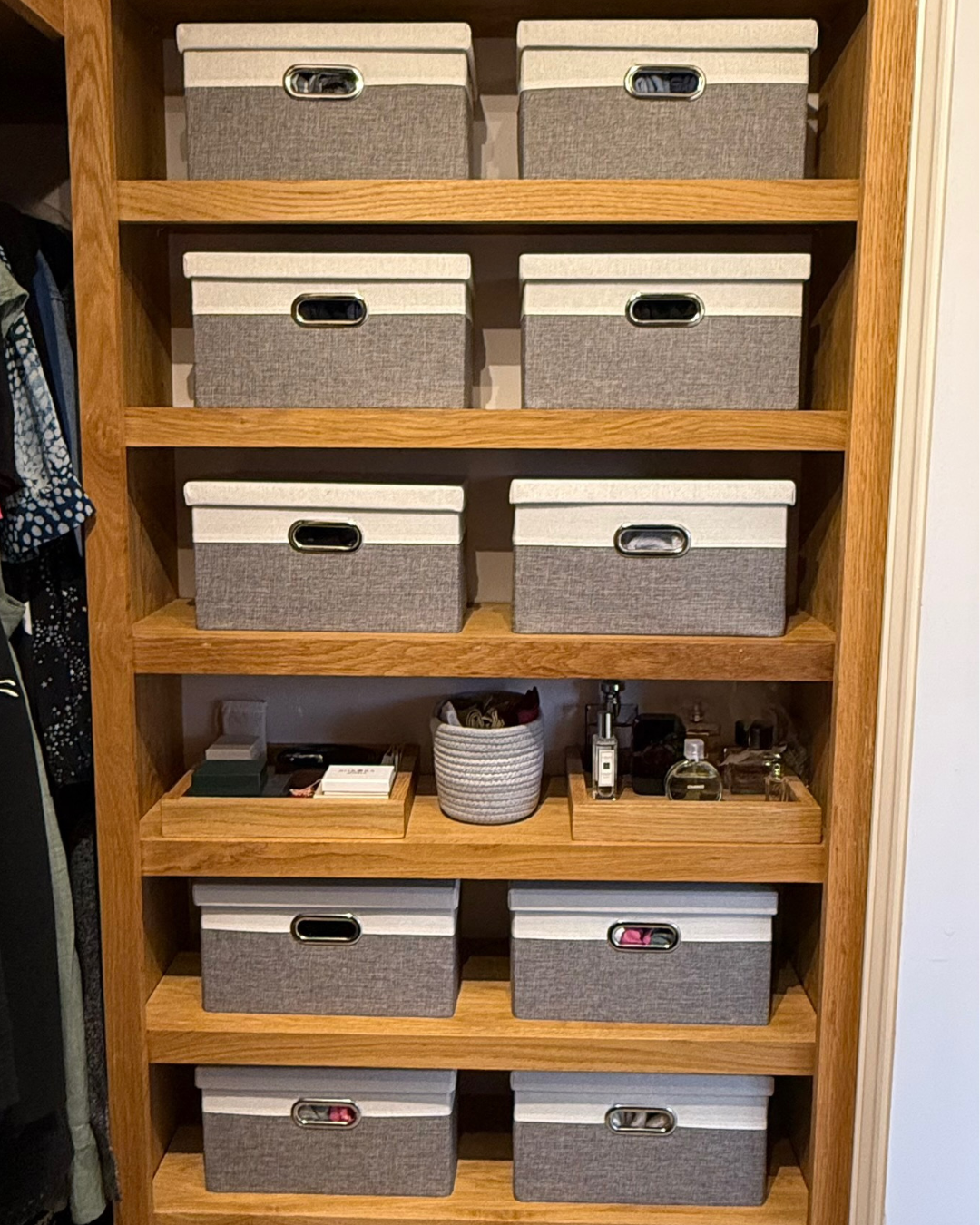 Wooden bookshelf with six shelves, filled with gray fabric storage bins on the top four shelves and small boxes, a plant, and perfume bottles on the lower shelf.