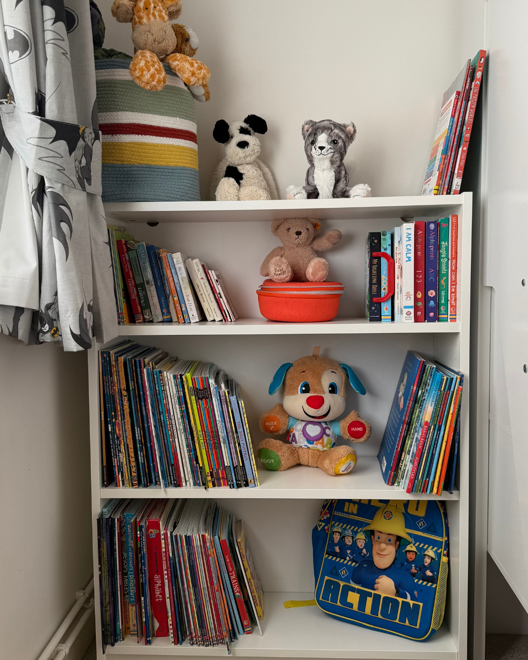 White bookshelf filled with children's books and plush toys, including a dog, bear, and firefighter backpack.