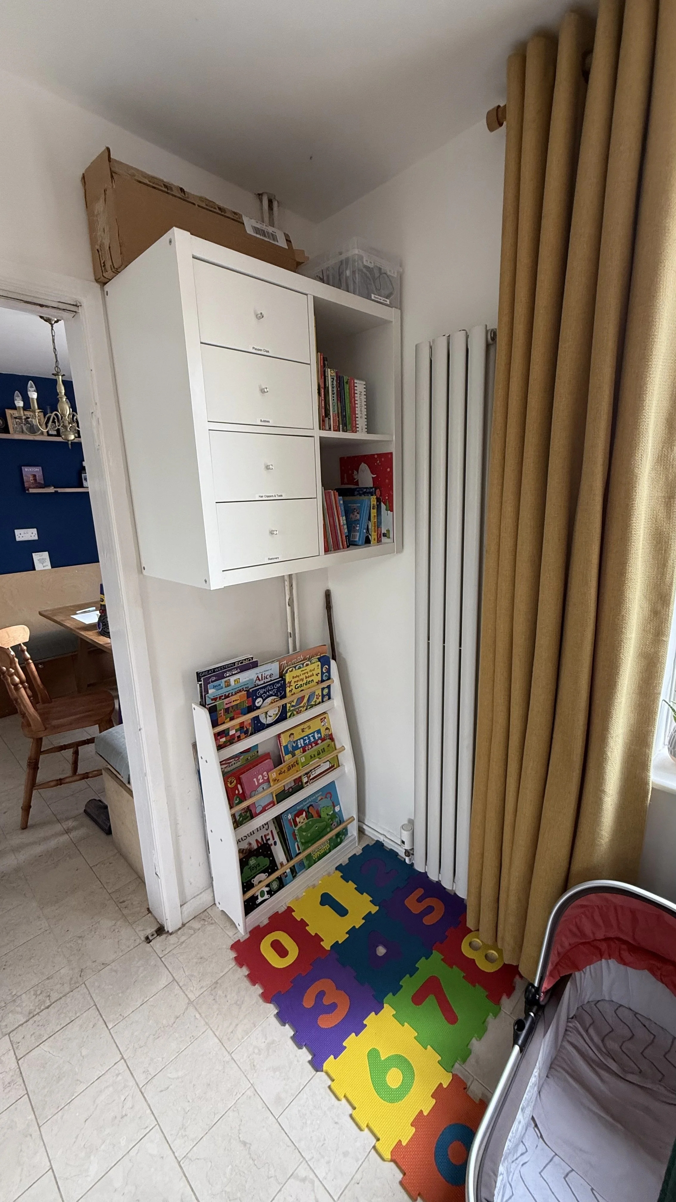 Children's area with a colorful foam alphabet and number rug, a bookshelf filled with children's books, a white wall-mounted mailbox with labeled drawers, and a toy storage rack with books. A portion of a baby crib is visible on the right, and beige 