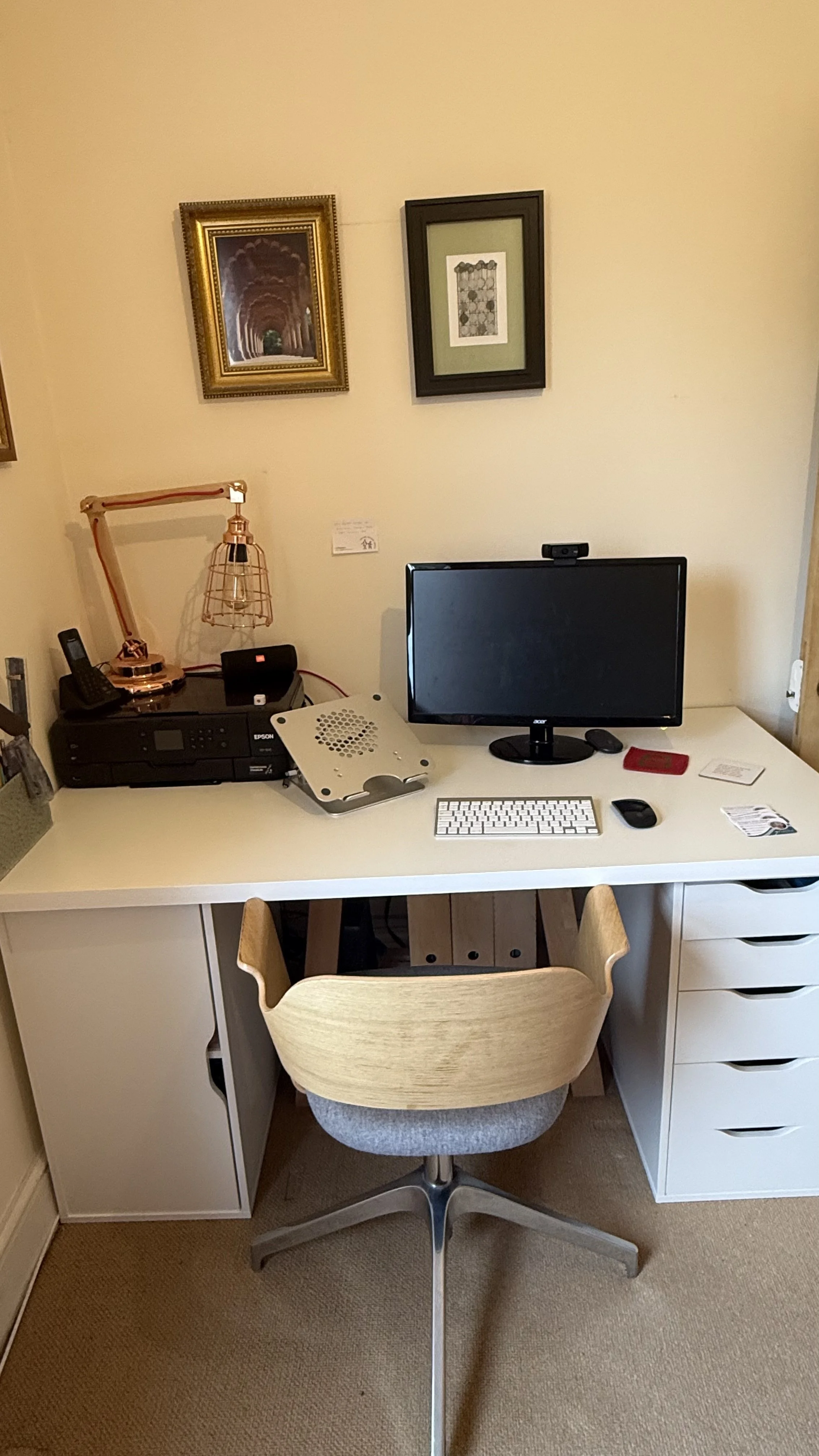 A tidy home office desk with a computer monitor, wireless keyboard, mouse, printer, and desk lamp. Framed artwork hangs on the wall above the desk. There is a swivel chair at the desk, set against a beige carpet.