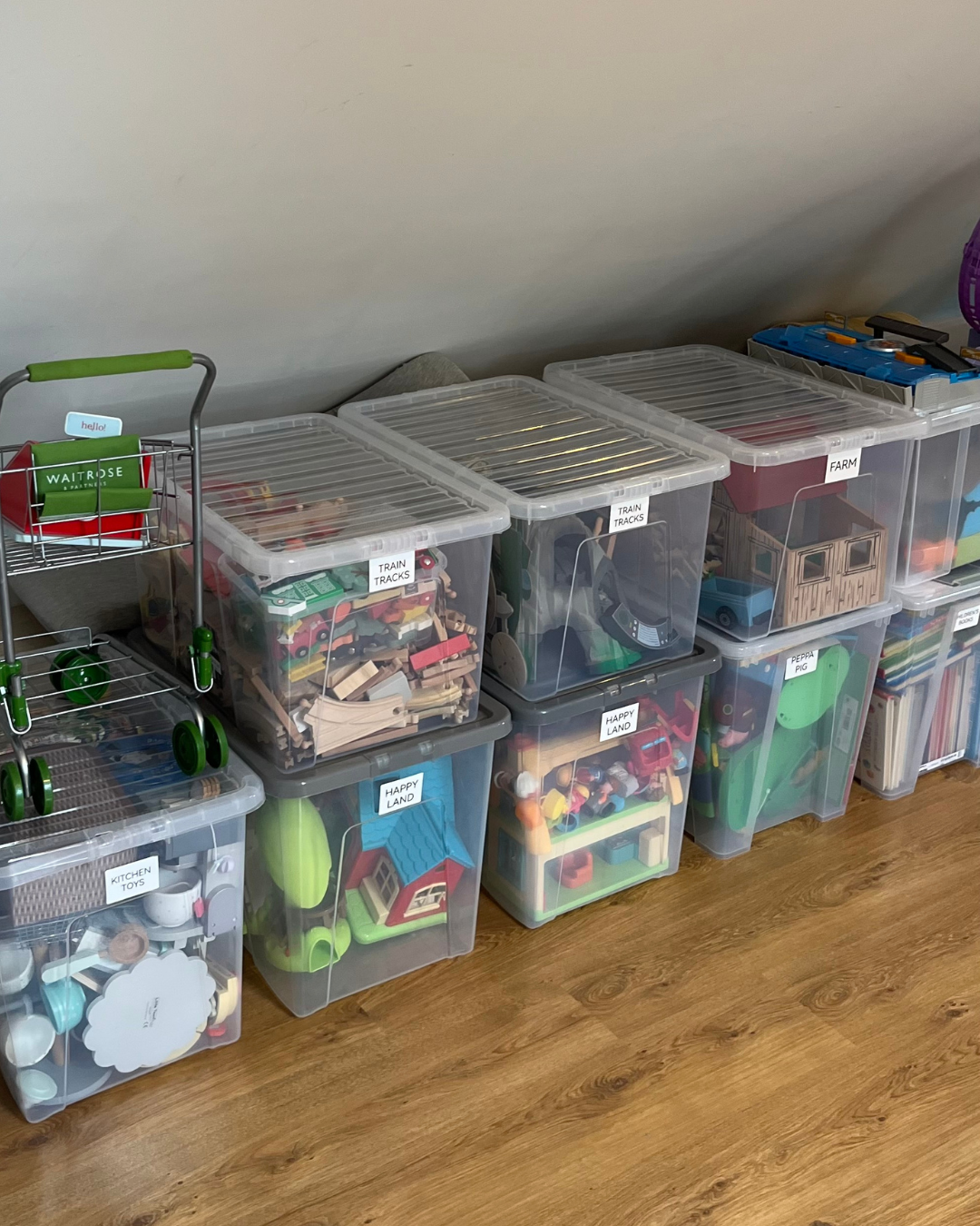 Storage bins filled with children's toys, labeled as train tracks, farm, happy land, kitchen toys, and peppa pig, organized on a hardwood floor against a wall.