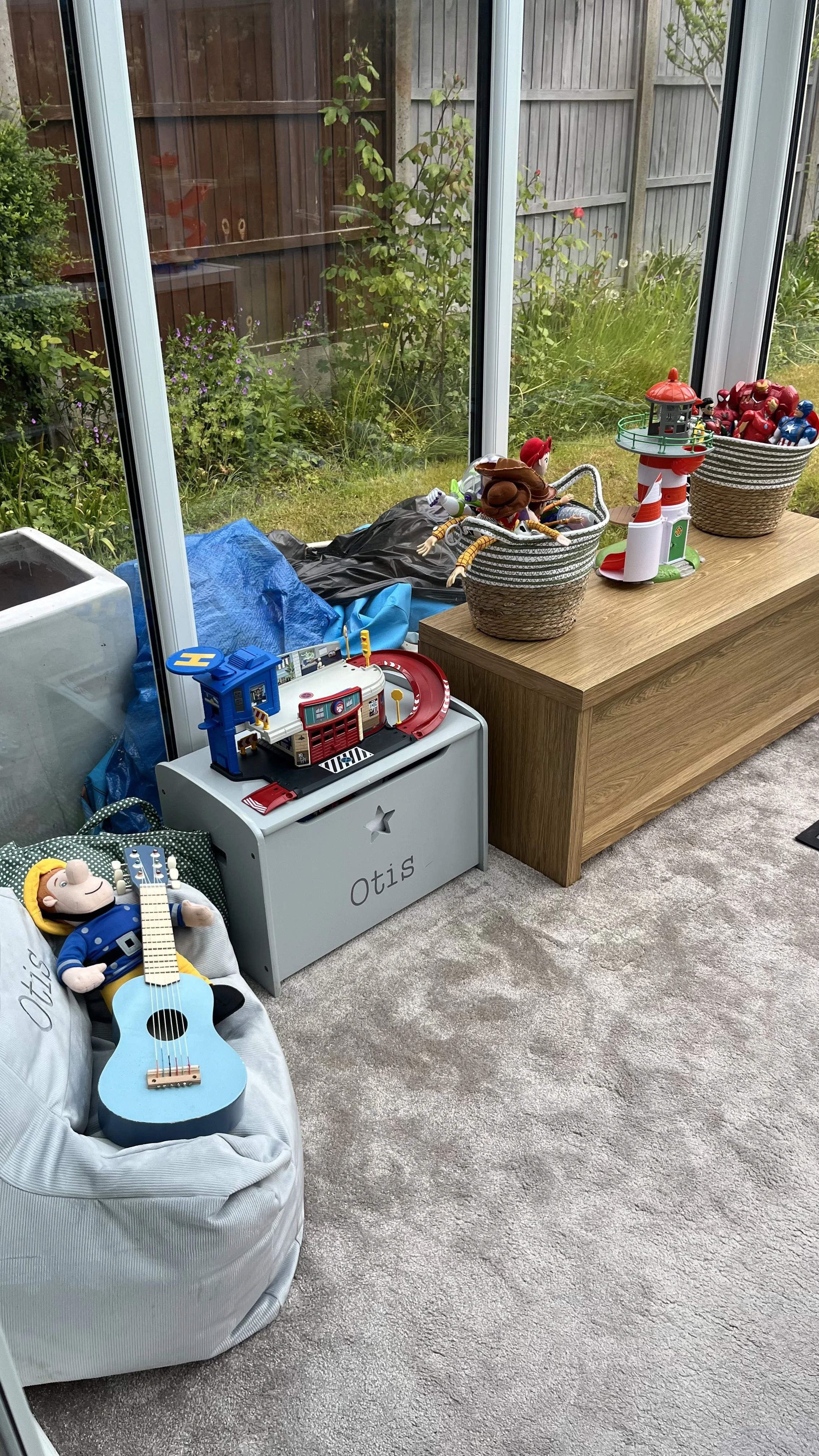 A playroom with toys, including a plush doll, a blue toy guitar, and various figures and playsets on and around a white storage box labeled "Otis." Behind the box, there is a wooden bench with baskets of toys and a view of a garden through large glass windows.
