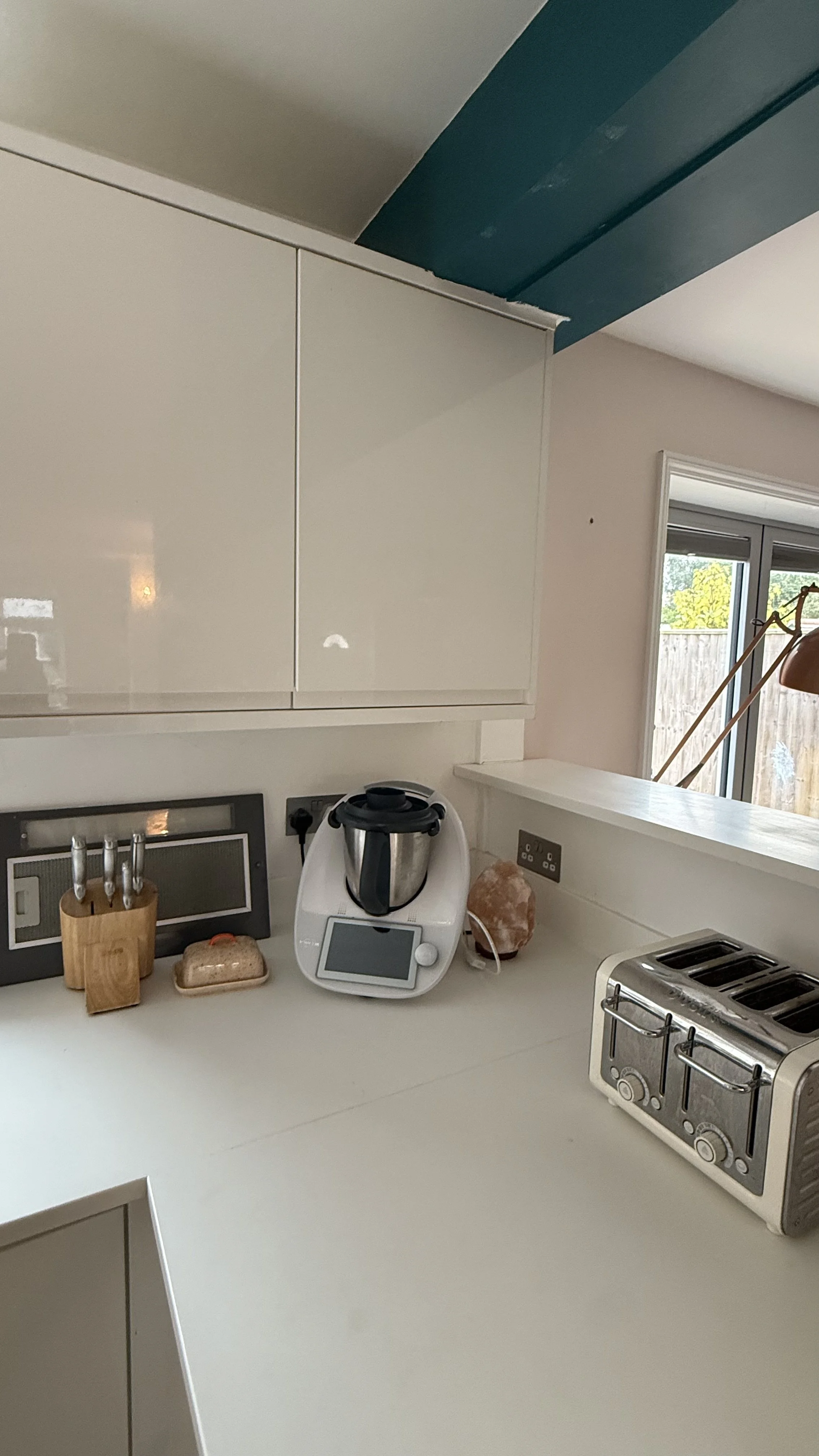Kitchen countertop with kitchen appliances including a multitask cooker, a four-slice toaster, a salt lamp, and a knife block. There is an upper white cabinet and a pillar of a blue ceiling beam.