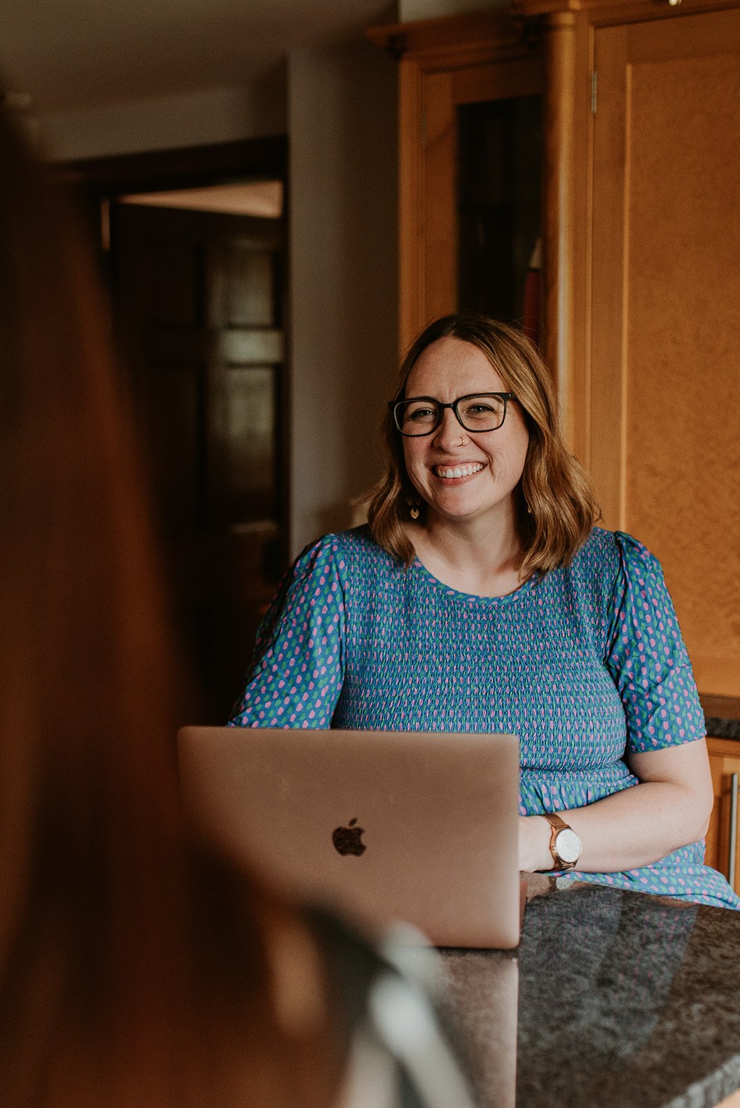 Smiling woman with glasses sitting at a kitchen table with a silver MacBook in front of her.