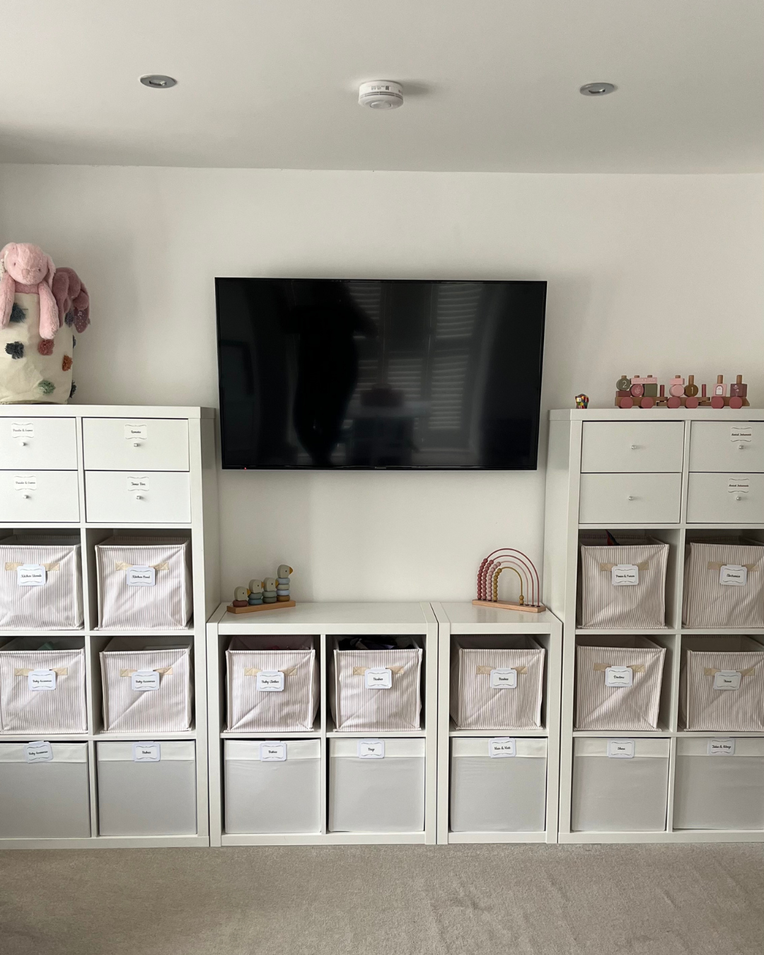 Living room with white storage units filled with labeled fabric bins, a large flat-screen TV on the wall, and children's toys including a stuffed animal and wooden rainbow decor.