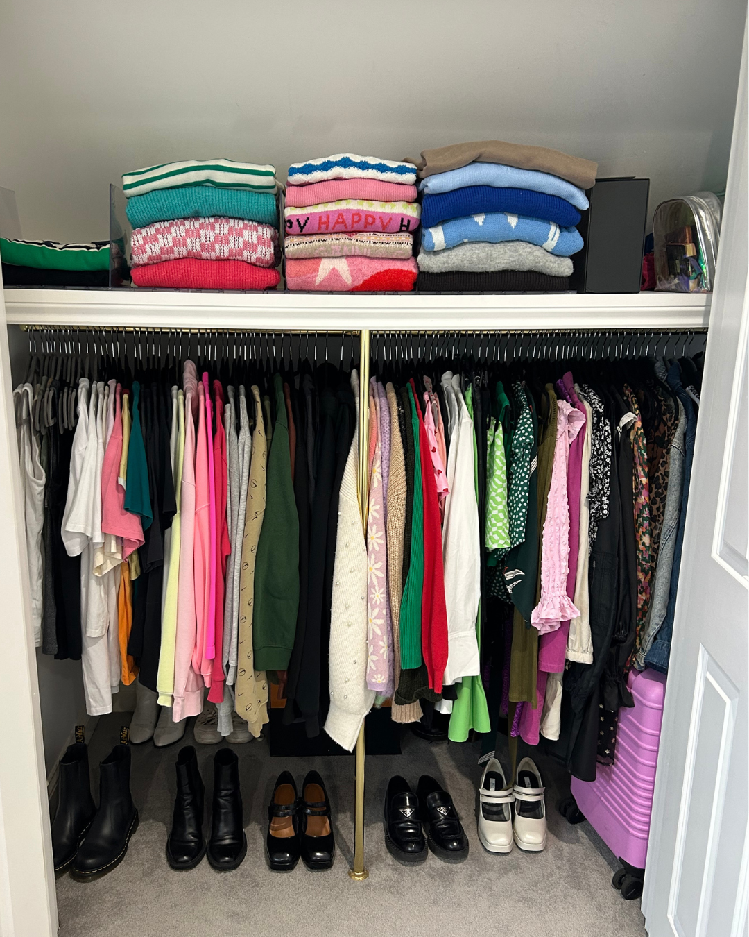 Closet with hanging clothes including shirts, sweaters, and dresses, along with folded sweaters and a pink suitcase on the top shelf, and several pairs of black and white boots and shoes at the bottom.