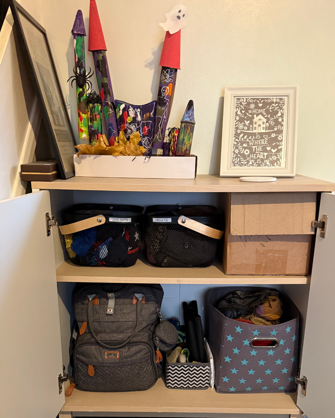 Open cabinet with two shelves filled with various bags, boxes, and craft supplies. Top shelf has labeled baskets, a framed quote, and a craft project with colorful paper cones and a ghost decoration. The lower shelf contains backpacks, a small box, a