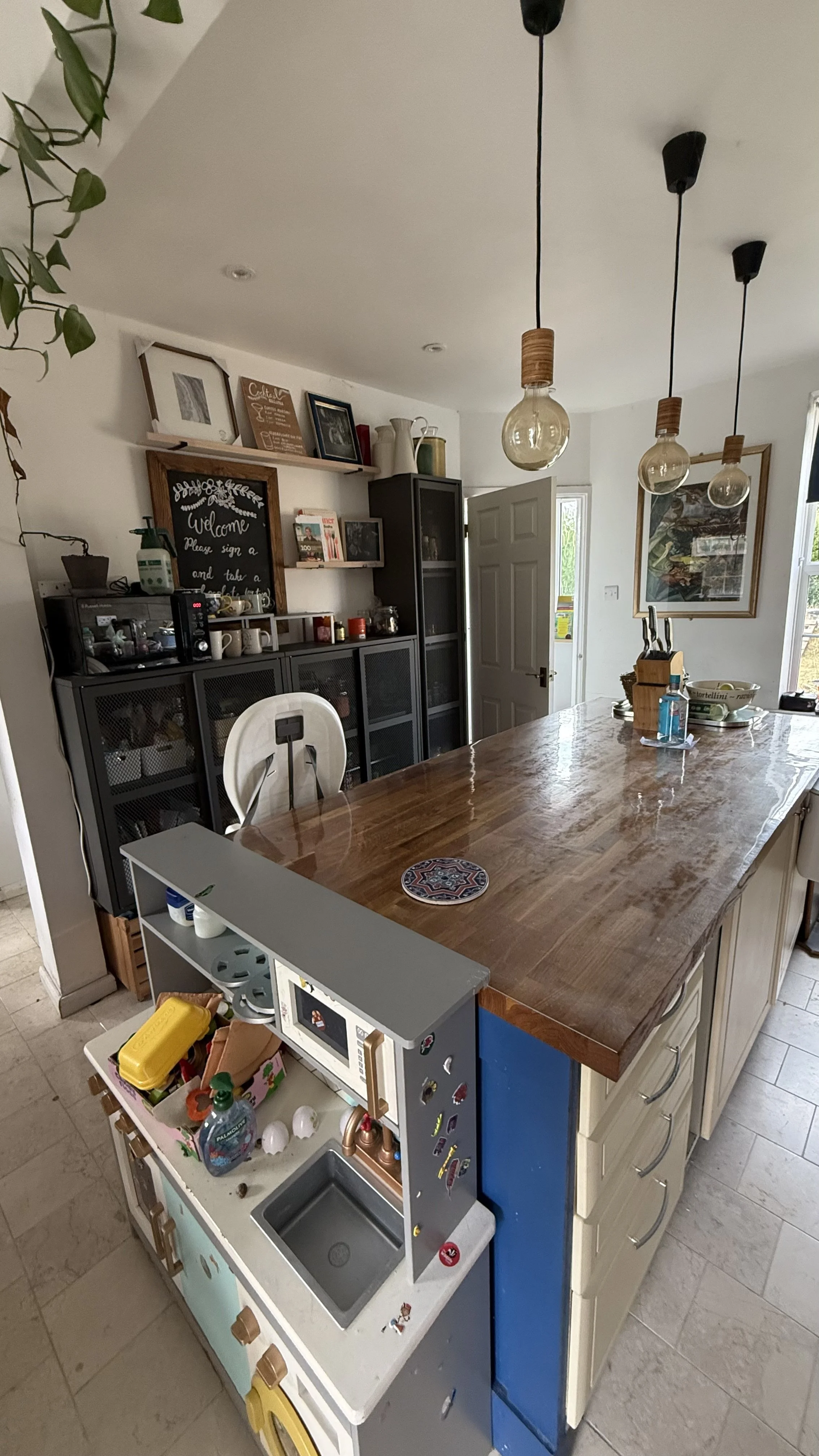 View of a kitchen and dining area with a large wooden table, hanging pendant lights, a black cabinet with glass doors, shelves with decor, a framed picture, and a small play kitchen set with toys.