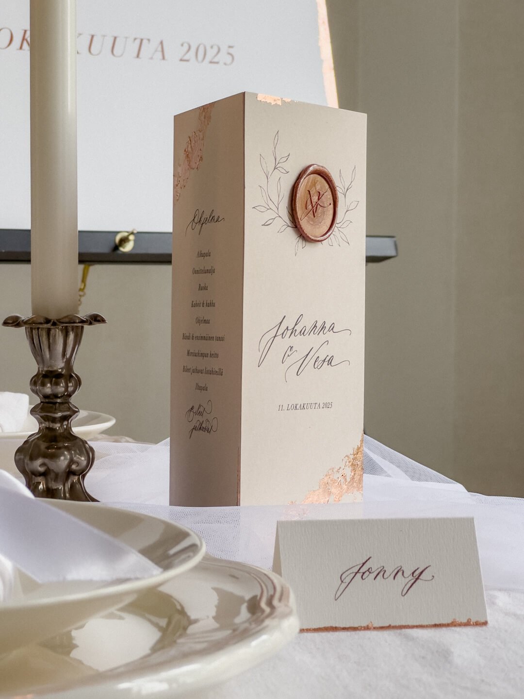 Syyshäiden kattauksen romanttiset paperituotteet. Pöydällä on pöytäkolmio, jossa on monogrammisinetti, kranssikuvio, lehtikultaa ja hääparin nimet kalligrafialla.