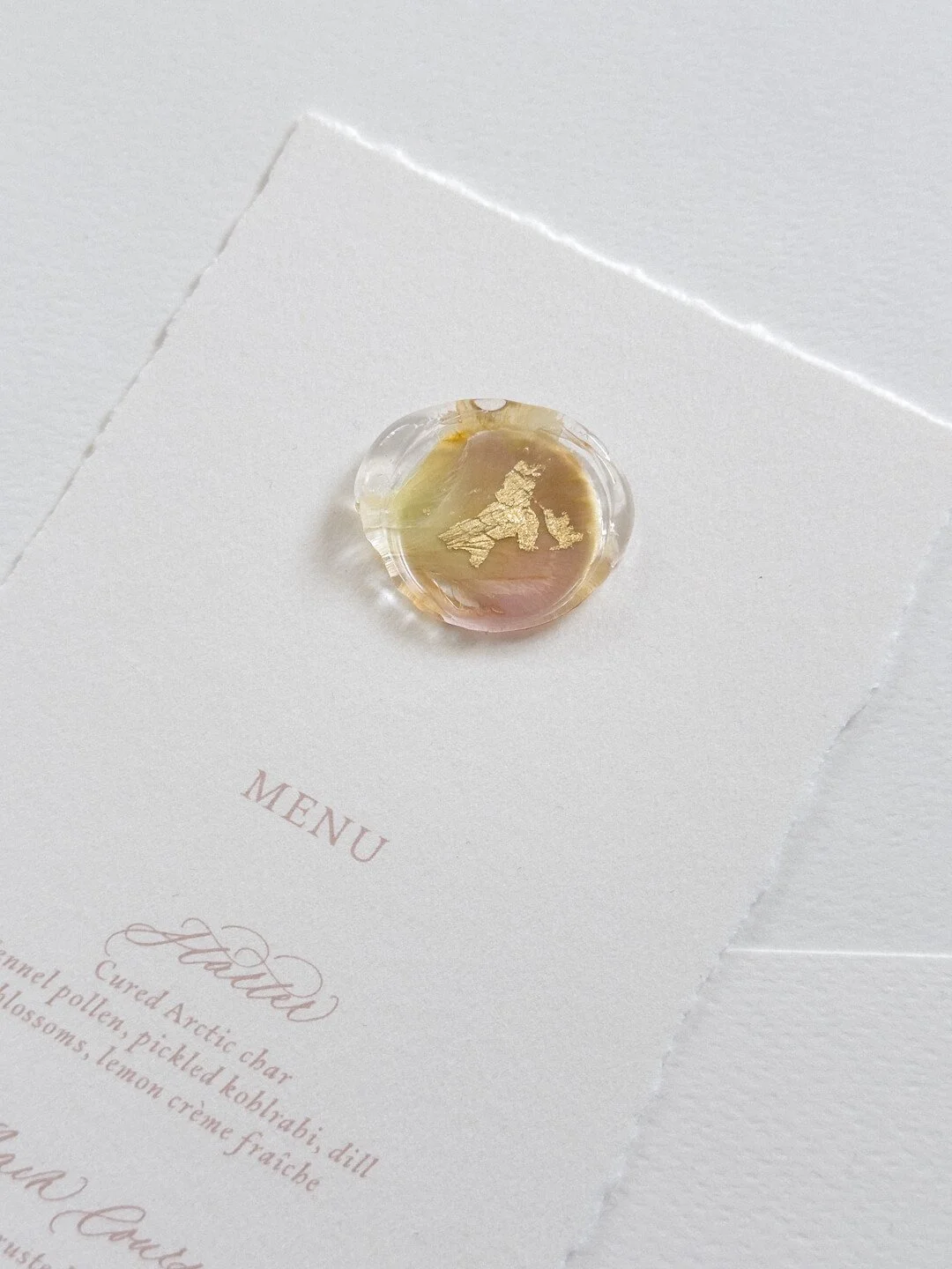 Uniikki kirkas sinetti häiden valko-vaaleanpunaisessa menukortissa. Sinetissä on kaunis, kuivattu kukka ja lehtikultaa.