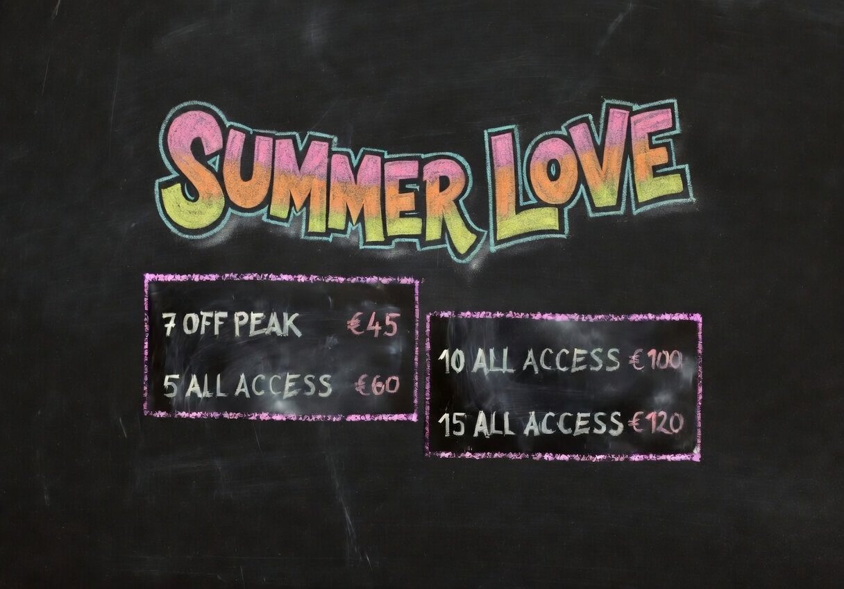 A chalkboard with colorful lettering that says 'SUMMER LOVE'. Below, there are four pricing options in two boxes: '7 OFF PEAK €45', '5 ALL ACCESS €60', '10 ALL ACCESS €100', and '15 ALL ACCESS €120'.