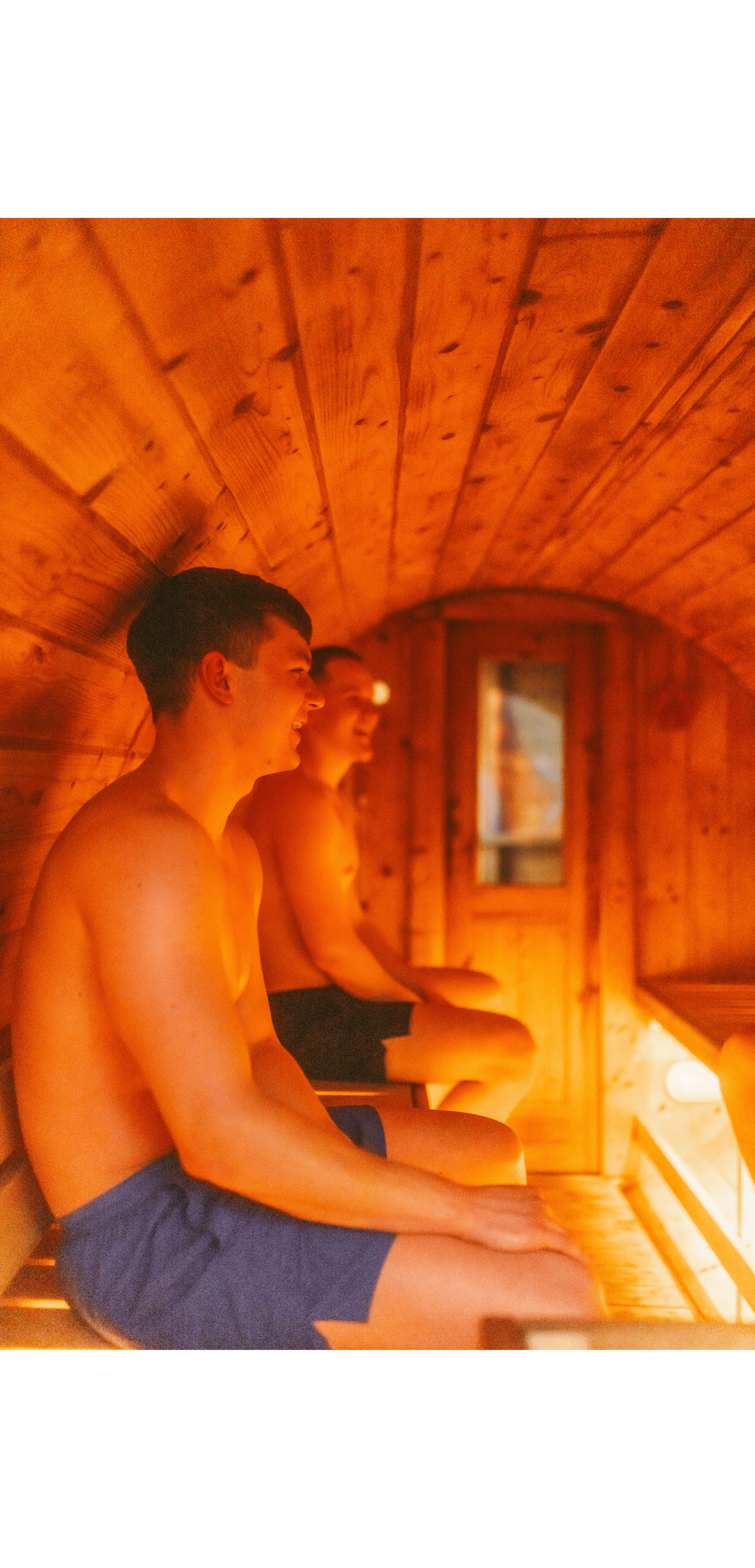 Two young men sitting inside a wooden sauna, smiling and relaxing, wearing swim trunks.