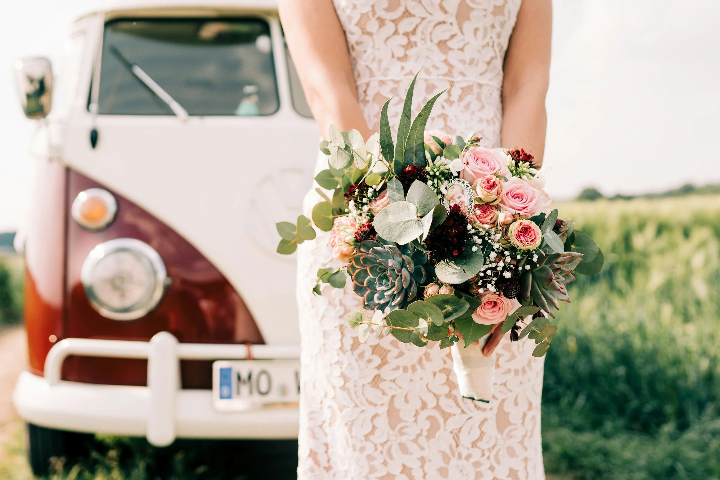 A woman in a light pink lace dress holding a bouquet of pink roses, greenery, and succulents, standing near a vintage white and maroon van.