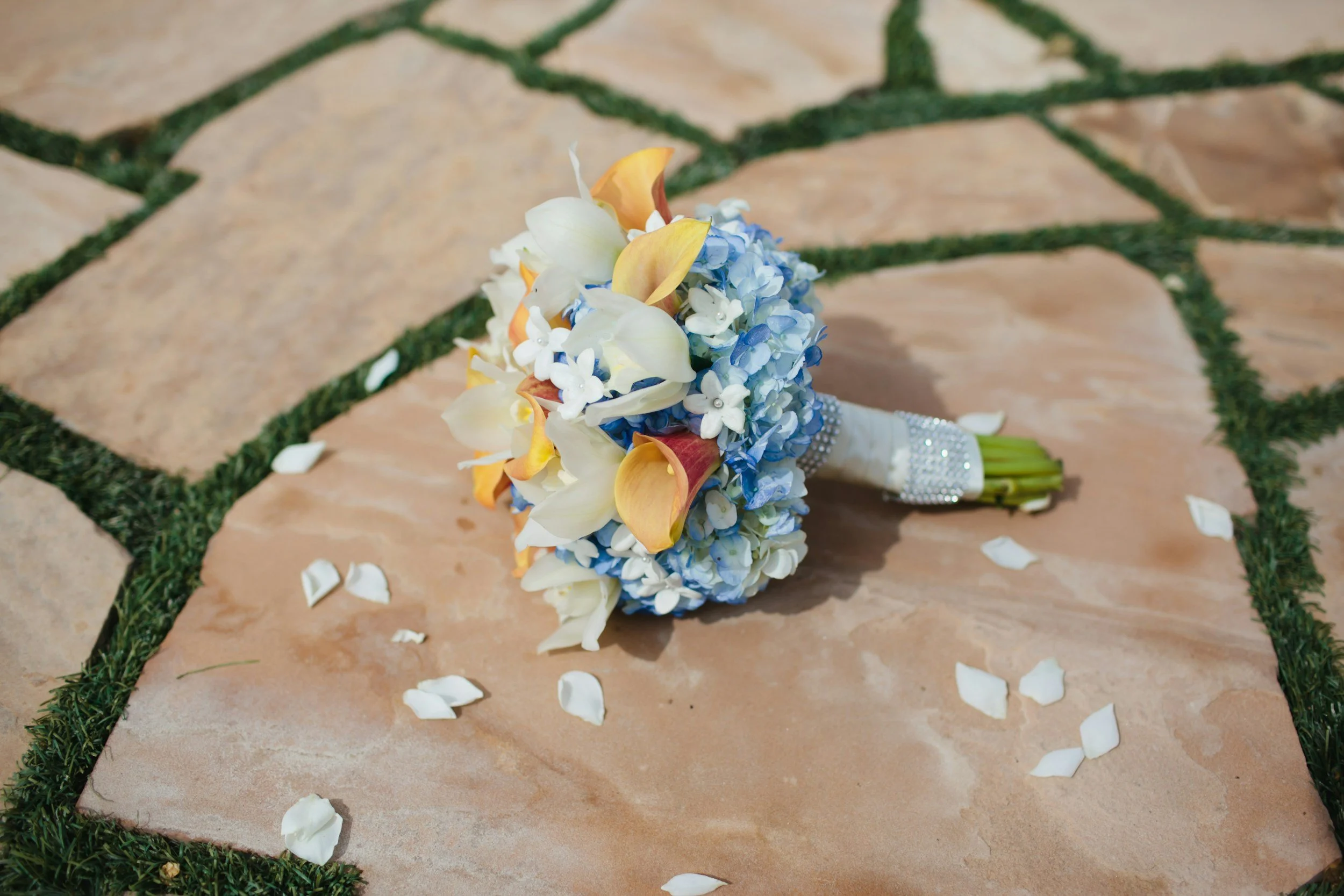 Wedding bouquet with white, yellow, and blue flowers resting on a tiled walkway with scattered white petals.