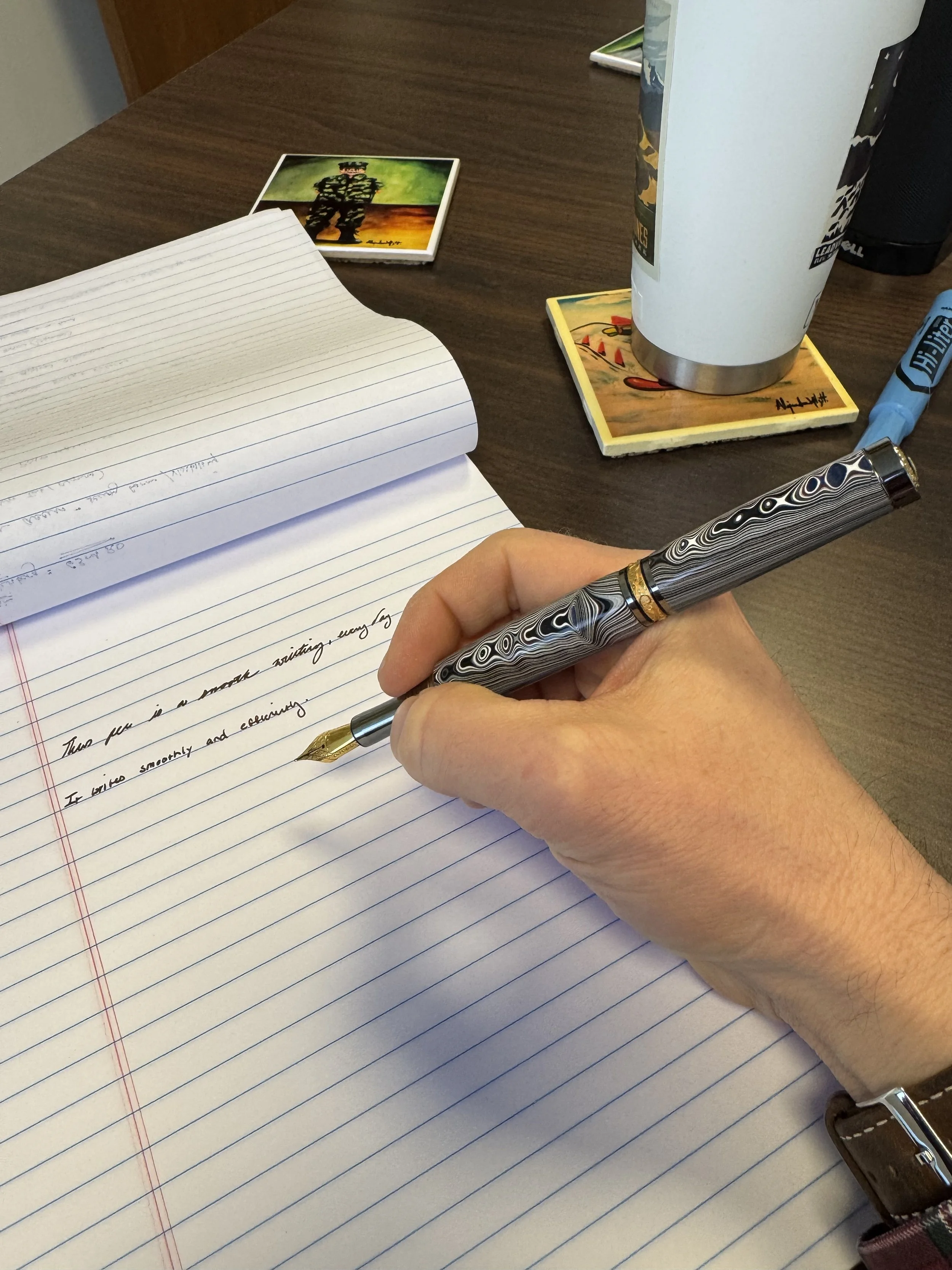 A person writing in a notebook with a fountain pen on lined paper at a desk. On the desk, there are two photographs, a large travel mug on a coaster, a blue highlighter, and a black reusable cup.