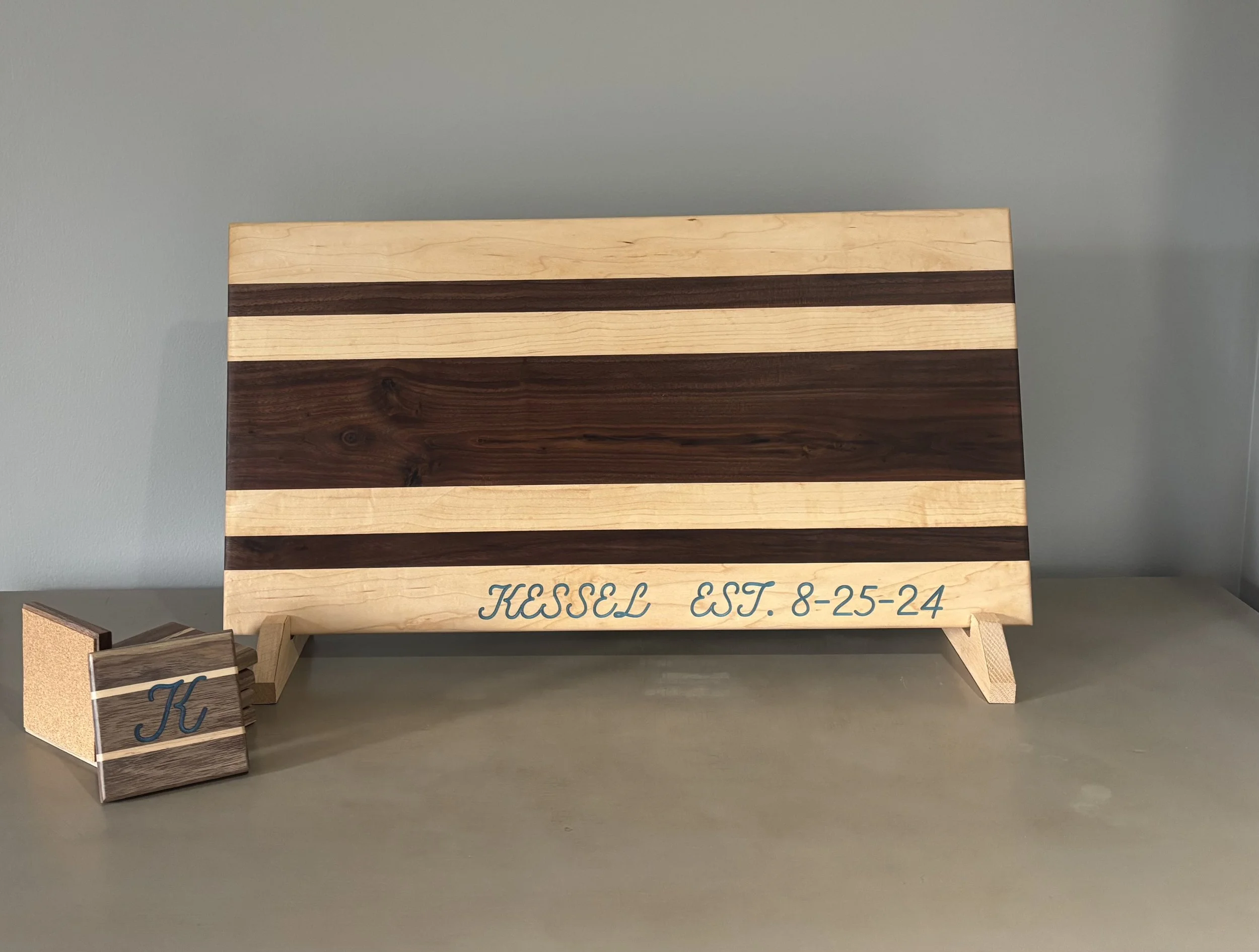 Wooden plaque with horizontal stripes in light and dark wood, inscribed with "Kessel EST. 8-25-24" in blue script, propped on small wooden stand with a smaller matching block with a blue letter "K" on it.