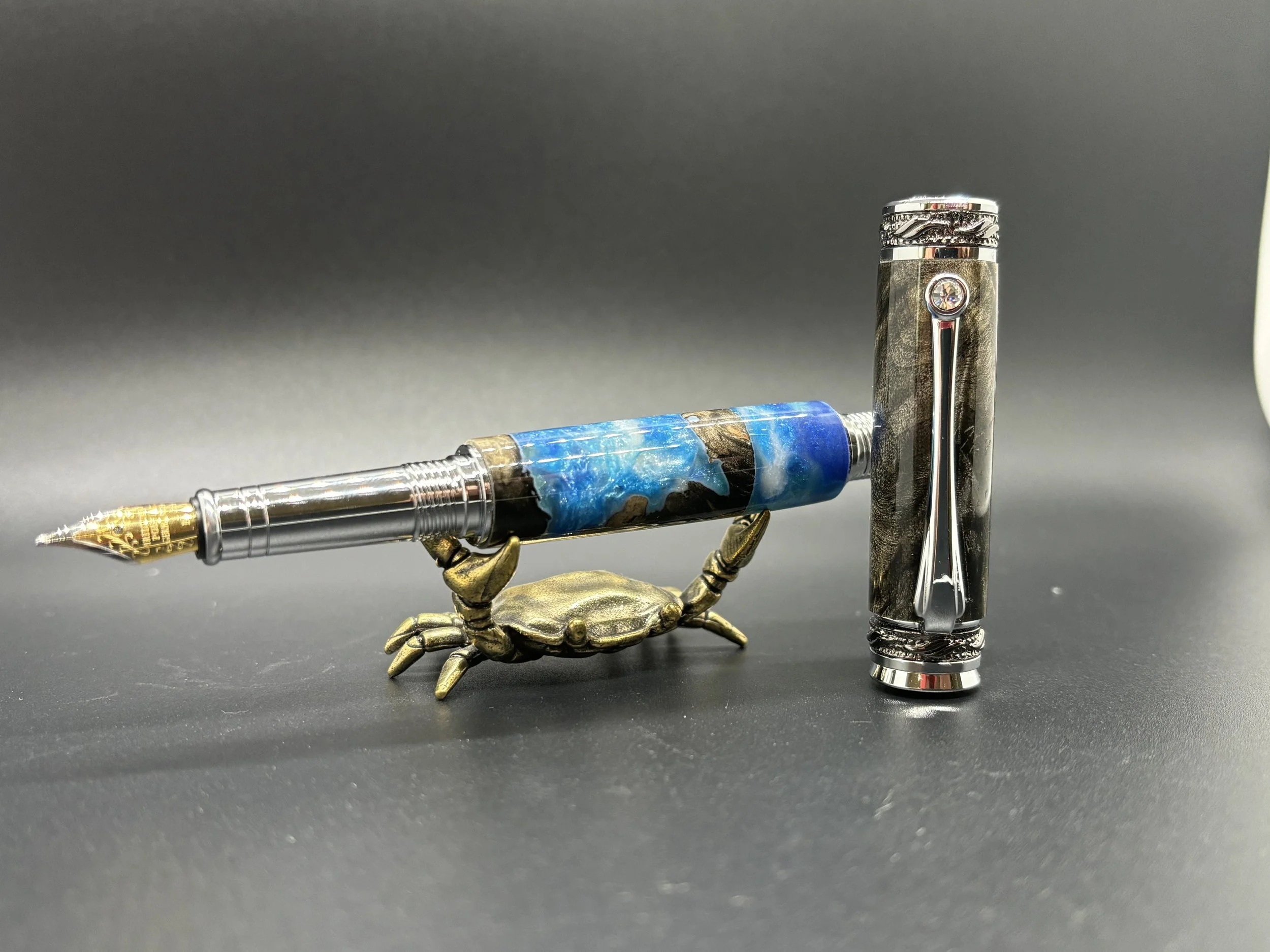 Decorative fountain pen resting on a brass crab figurine, with a black and silver fountain pen standing upright to the right.