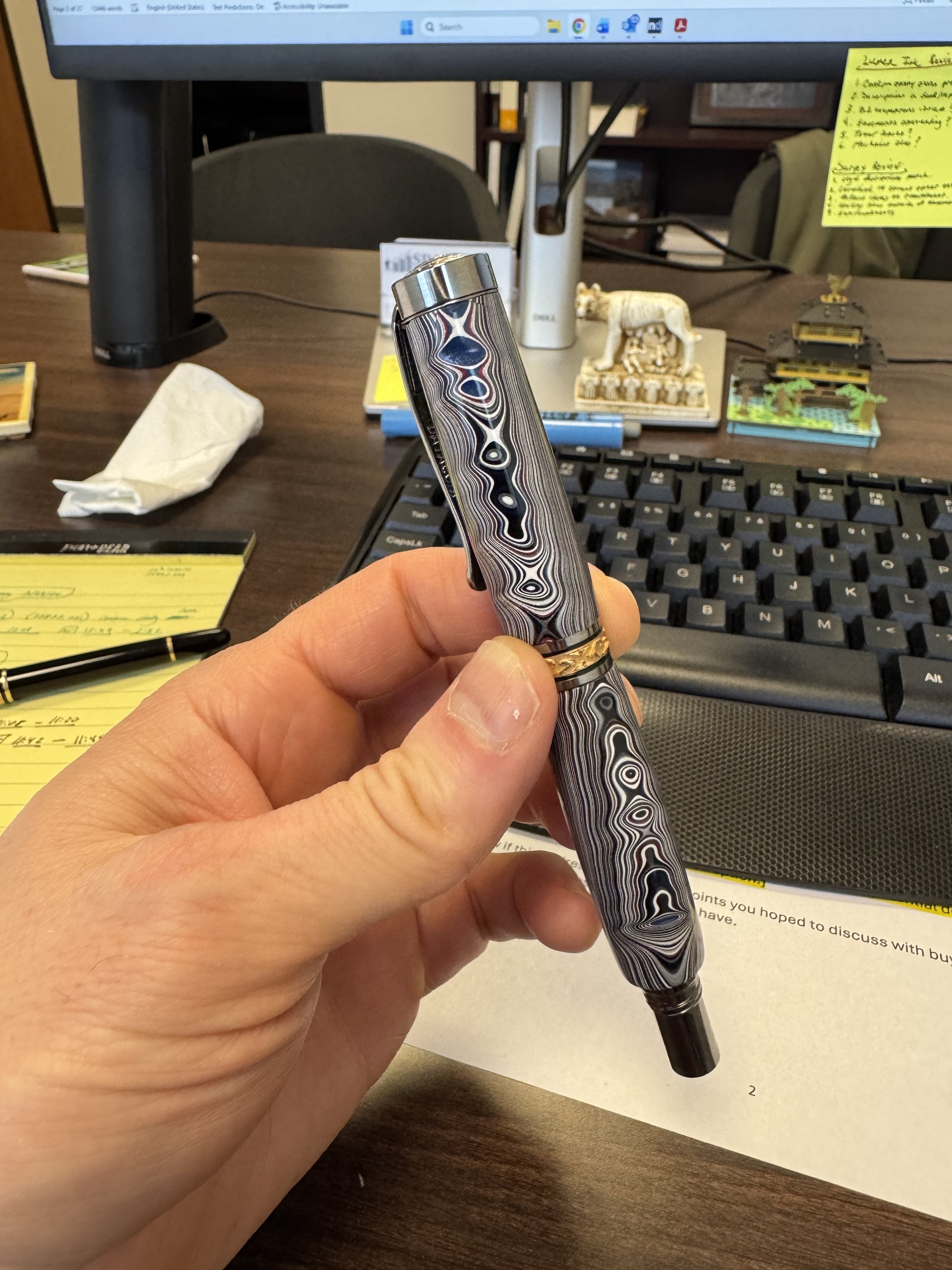 Hand holding a patterned pen with a swirling black, white, and gray design, gold ring near the middle, in an office setting with a computer keyboard, yellow notes, and various small decorative items on the desk.
