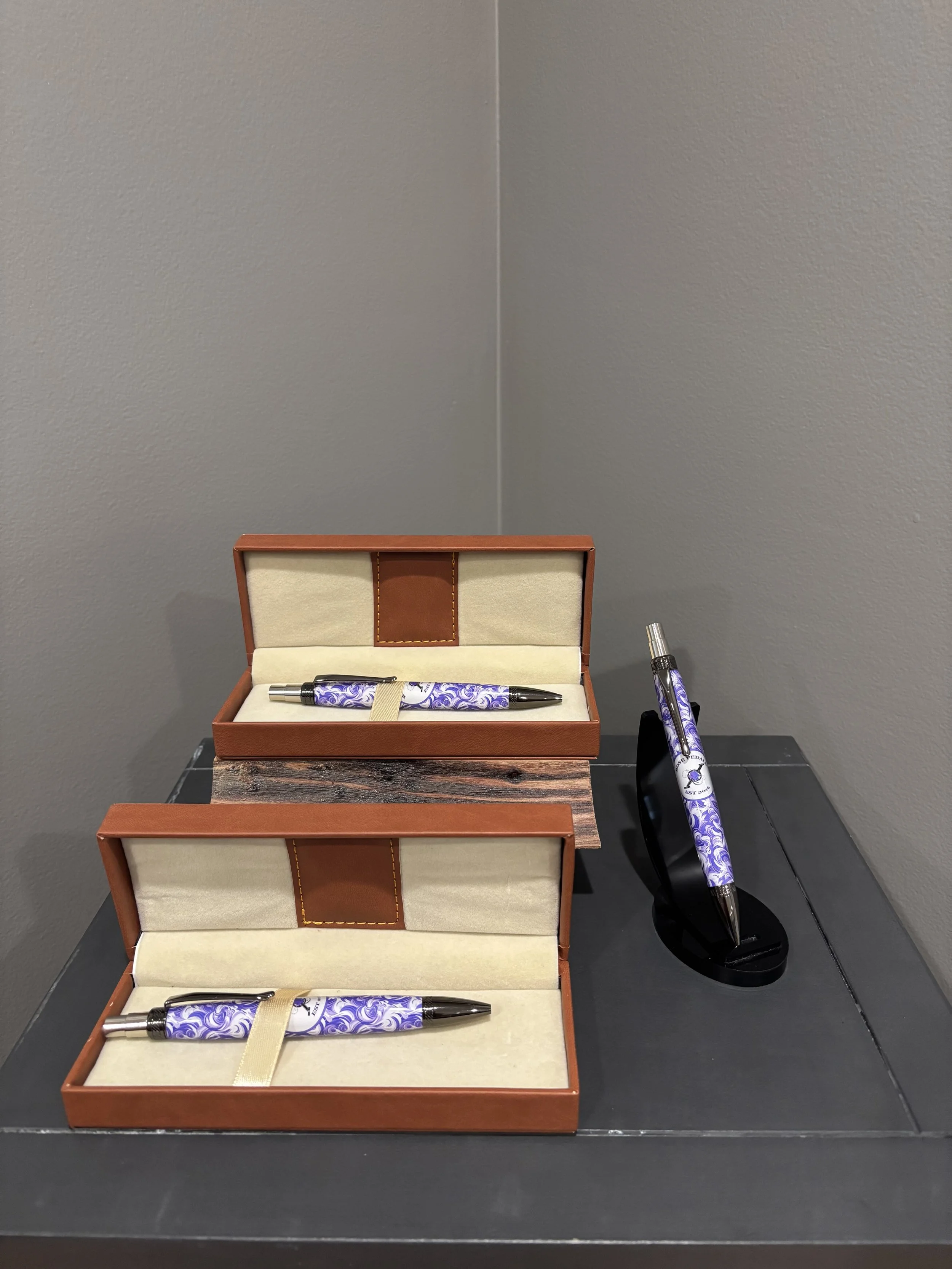 Two pens with purple and white swirling designs in brown leather boxes with cream interior, and a matching pen on a black stand, all placed on a black table against gray walls.
