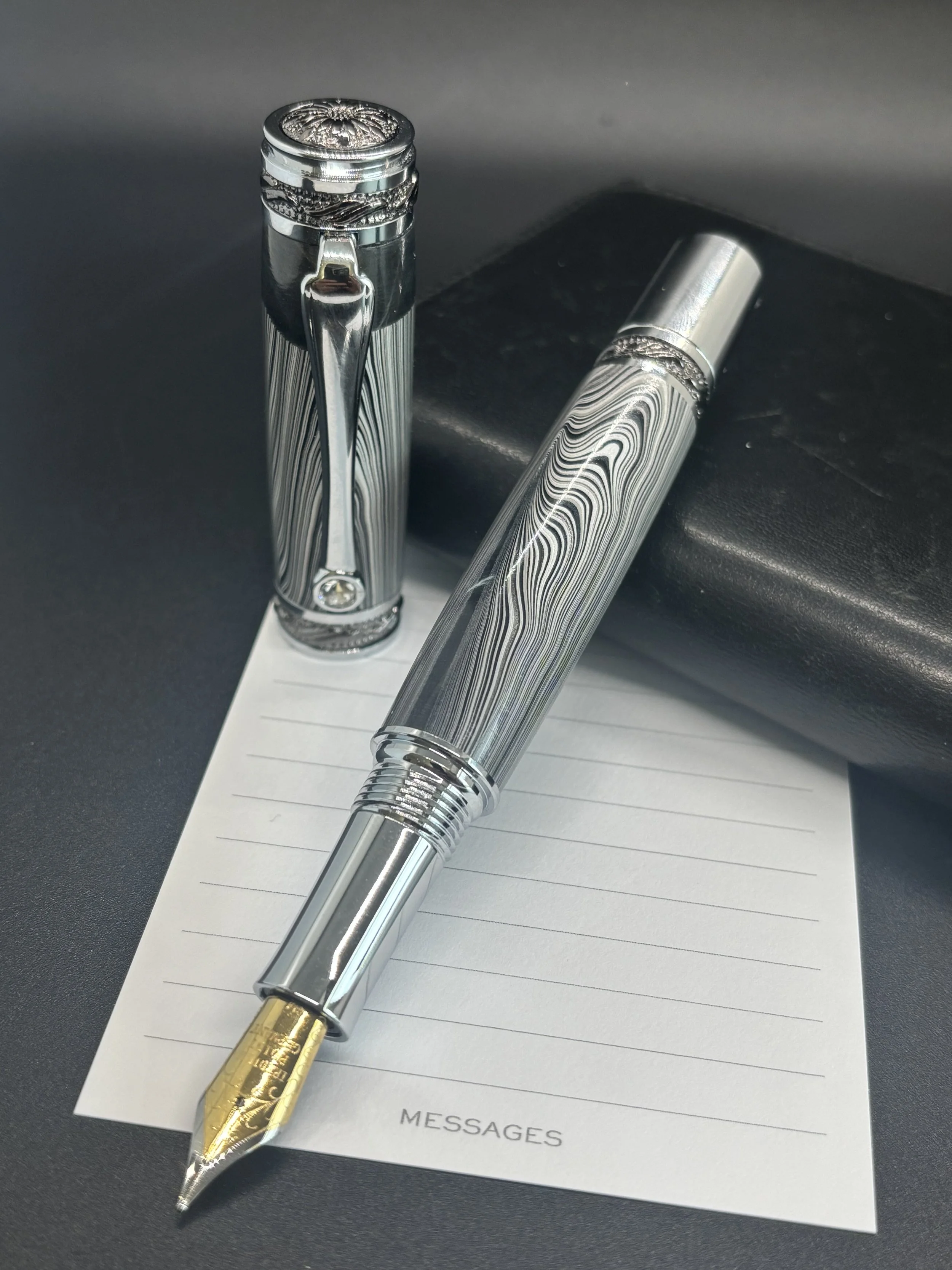 A silver fountain pen with a patterned body and gold nib, placed on a lined notepad labeled 'Messages', with a matching patterned cap resting nearby.