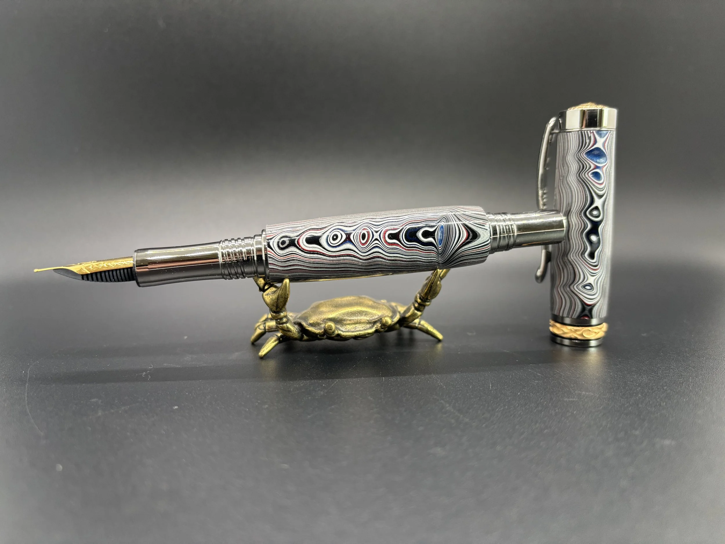 A decorative fountain pen with a swirling black, white, red, and blue pattern on its body and cap, placed on a black surface. The pen is supported by a small bronze-colored crab figurine.