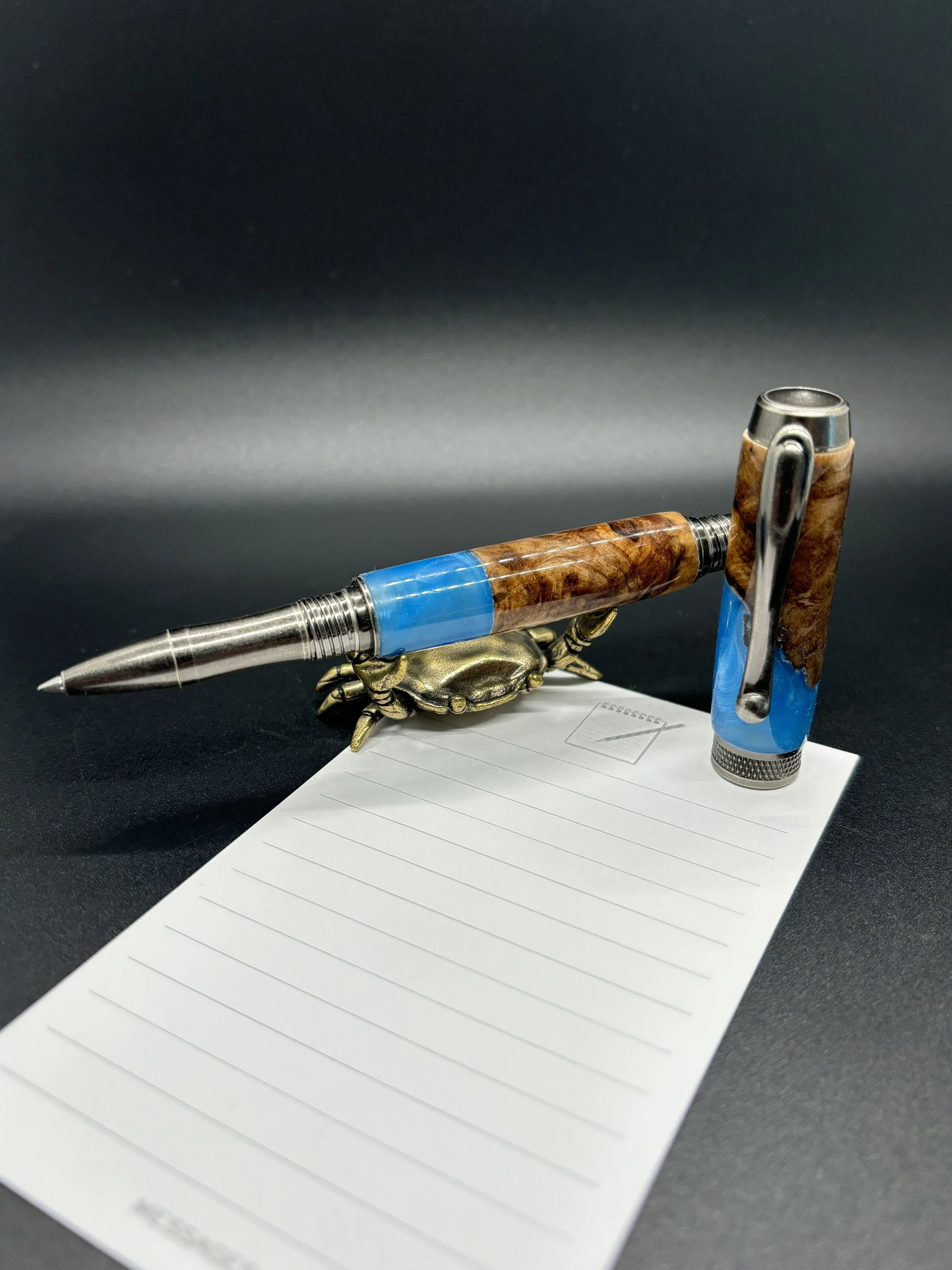 A decorative fountain pen with a wooden and blue resin body, resting on a small brass crab figurine, next to a lined notepad on a black surface with a dark background.