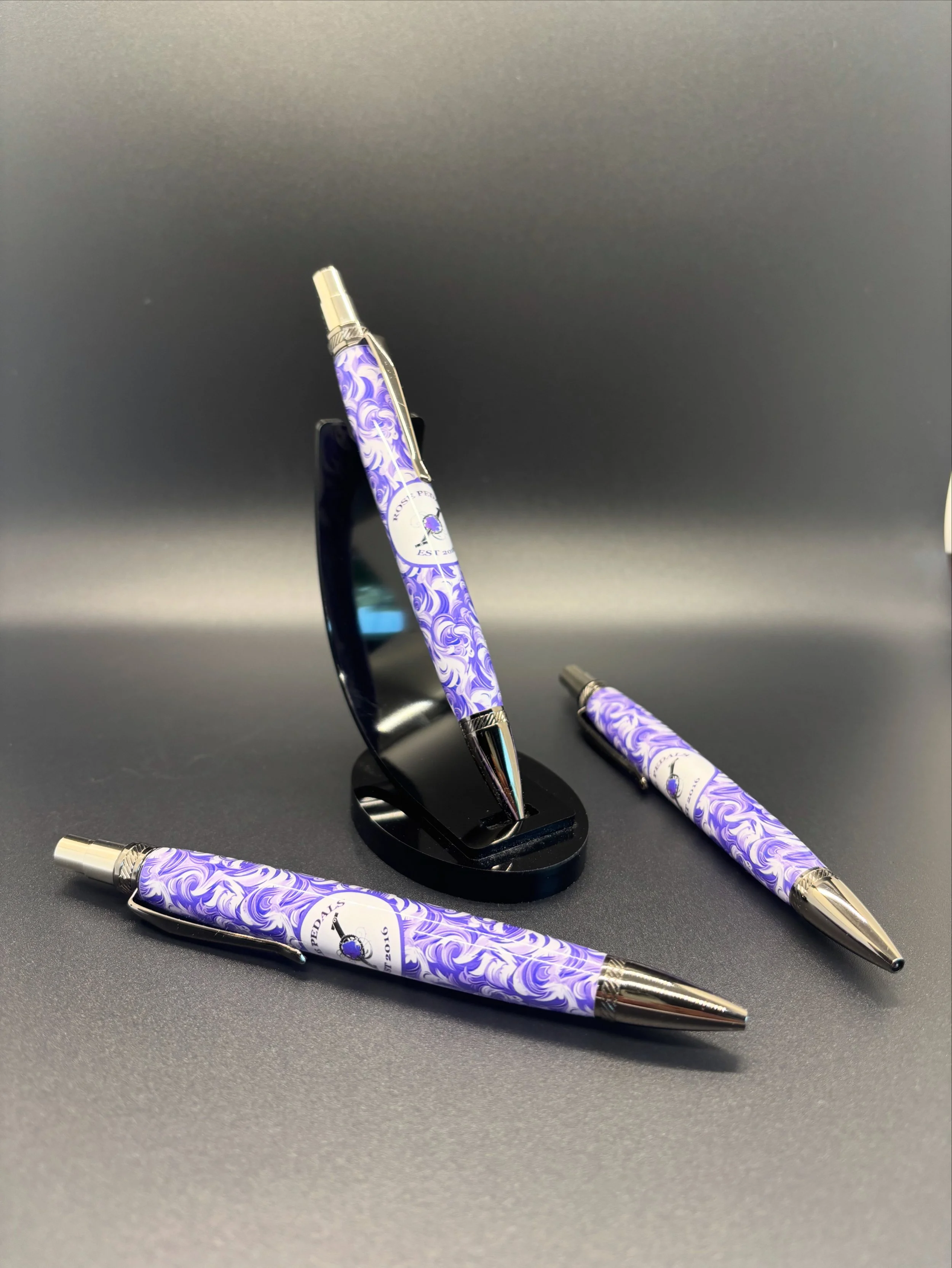 Set of three pens with purple swirling patterns and silver accents, one standing in a black pen holder and two laying flat on a dark gray surface.
