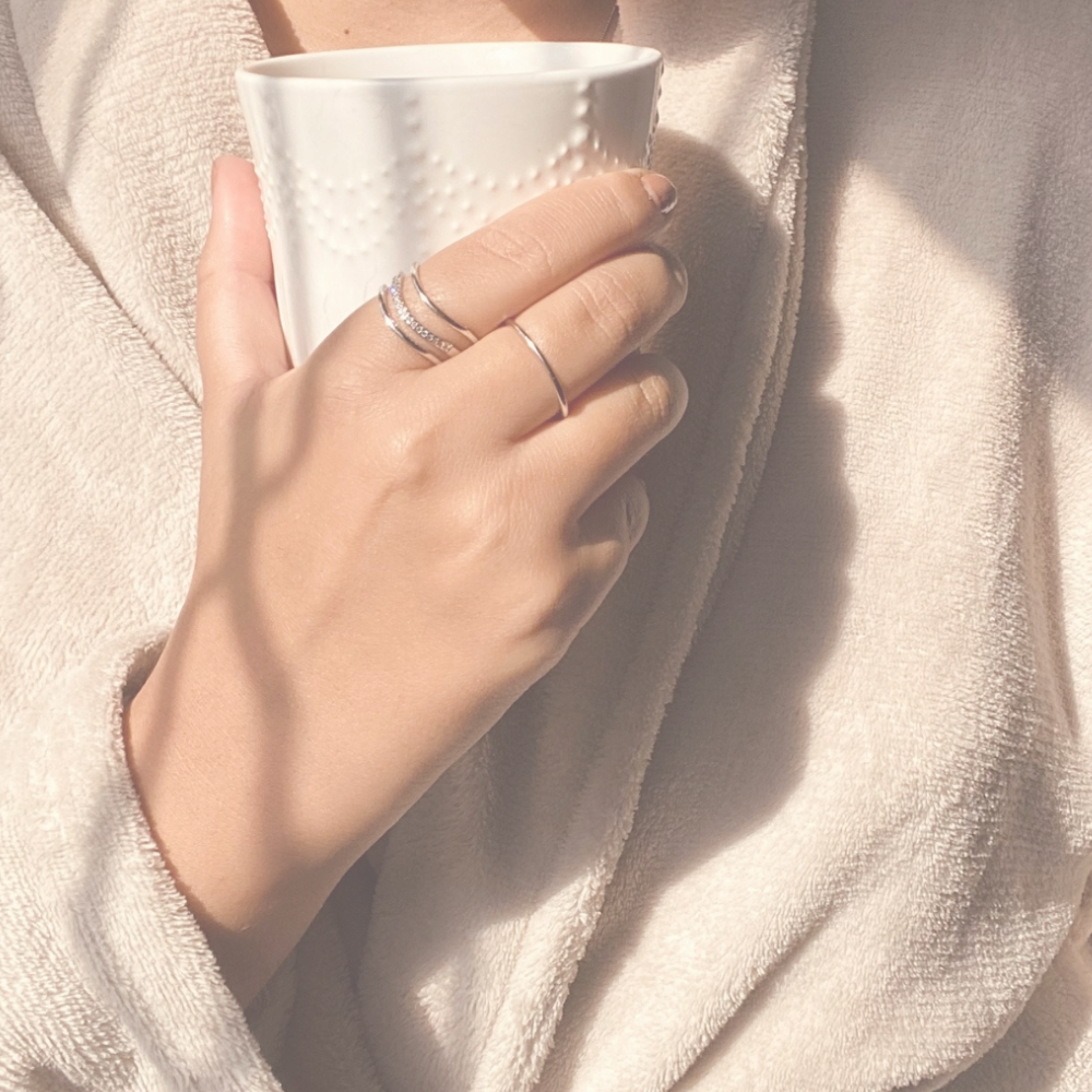 Person holding a white ceramic mug with textured design, wearing multiple silver rings, dressed in a light-colored, cozy robe.