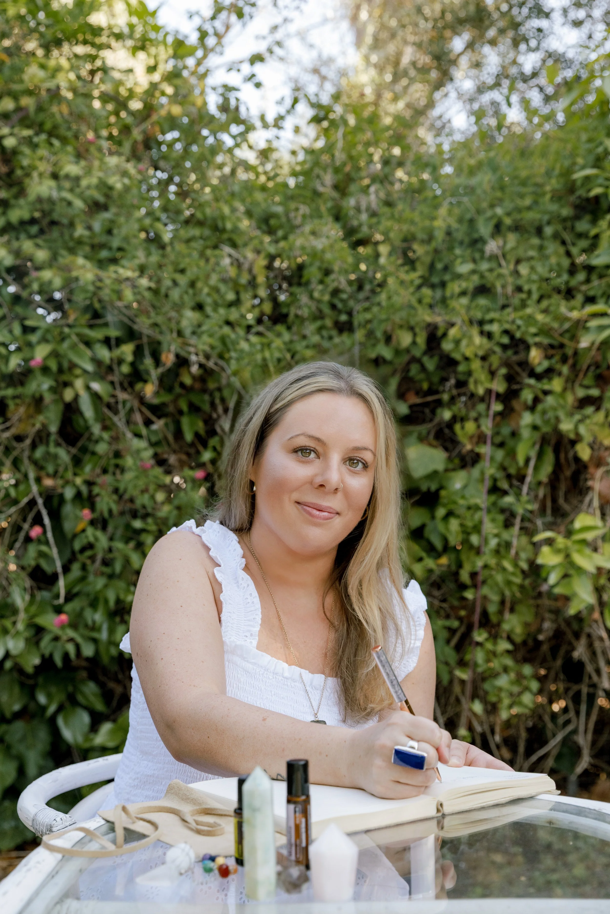 A woman with blonde hair and a septum piercing, wearing a white sleeveless top, is sitting at a table outdoors with a greenery background. She is holding a pen above an open notebook, surrounded by art supplies and bottles.