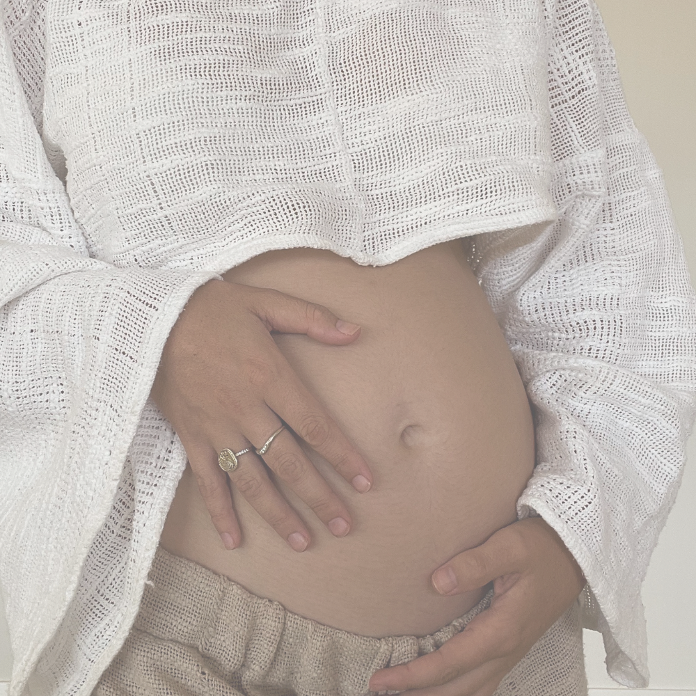 A pregnant person gently touching their belly with one hand and resting the other hand on top, wearing a white textured, open-knit top and beige shorts.
