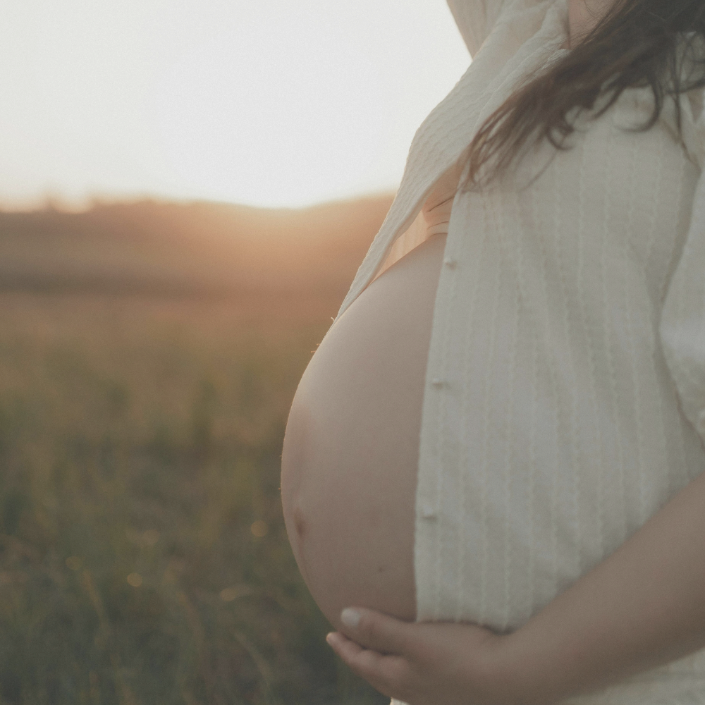 A pregnant woman outdoors at sunset, wearing a cream-colored textured shirt, with her hand resting on her pregnant belly.