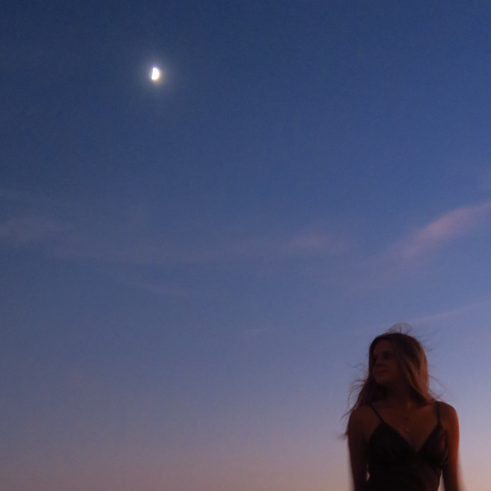 A woman with long hair in a black dress stands outdoors at dusk with a dark blue sky and a crescent moon overhead.